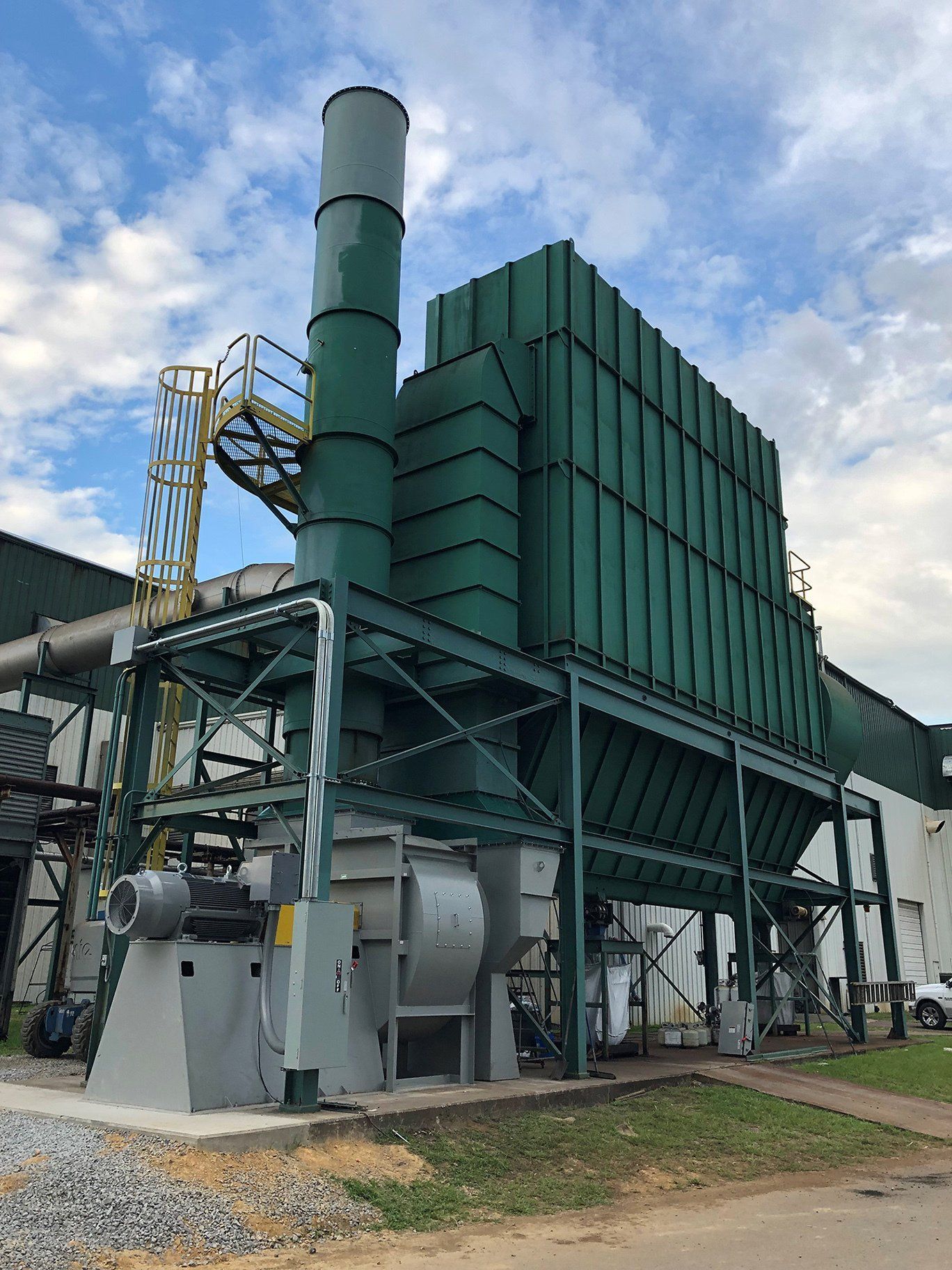 Green industrial dust collection system with a tall chimney, beside a building.