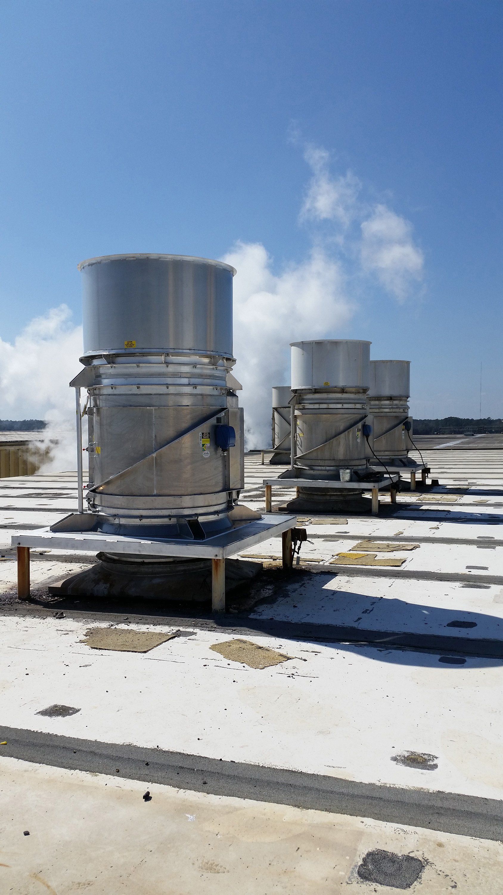 Rooftop HVAC units venting steam into a blue sky.  Silver cylinders on a white surface.