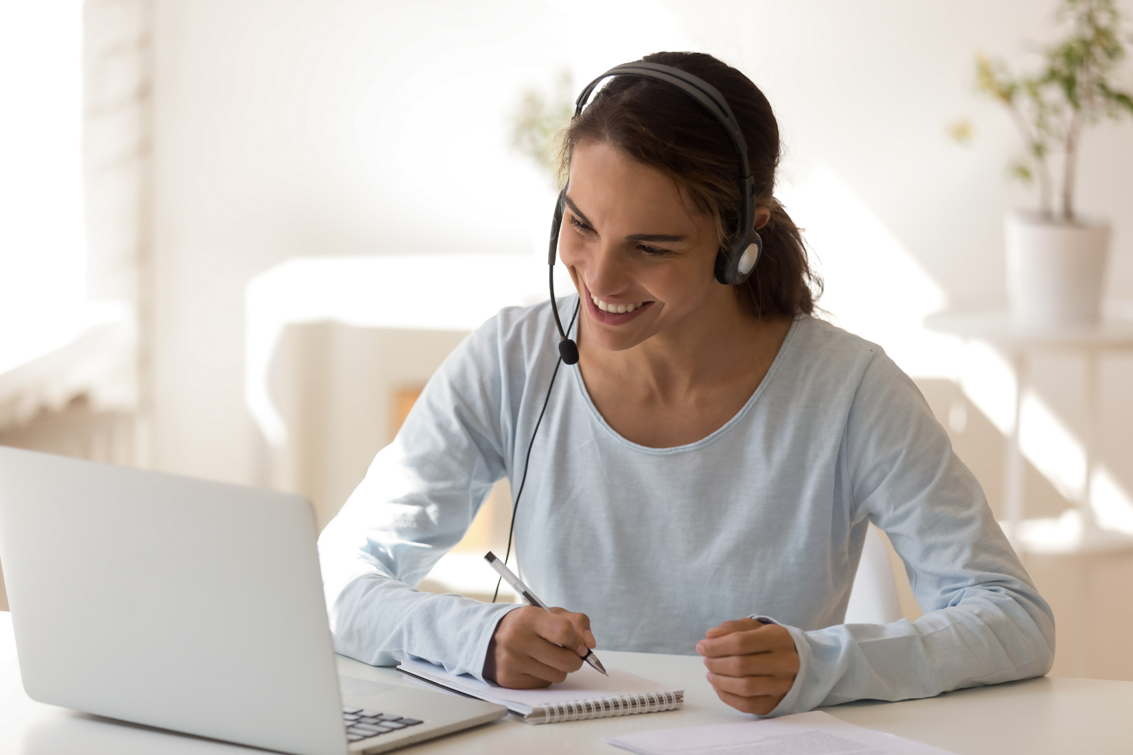 Smiling person wearing headphones writes in a notebook beside a laptop at a bright desk