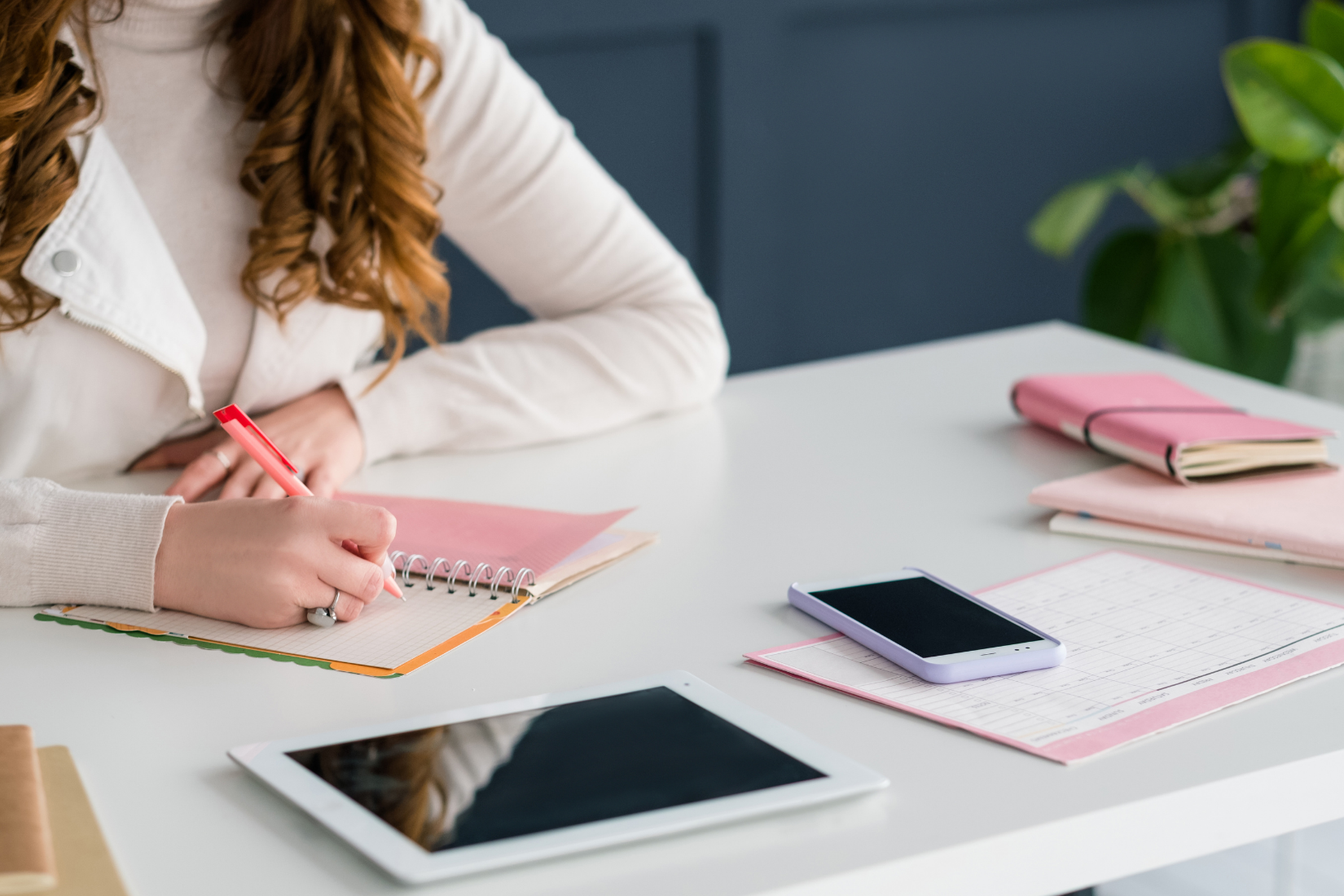 Woman writes in a notebook at a white desk. Pink notebook, smartphone, tablet, and journal visible.