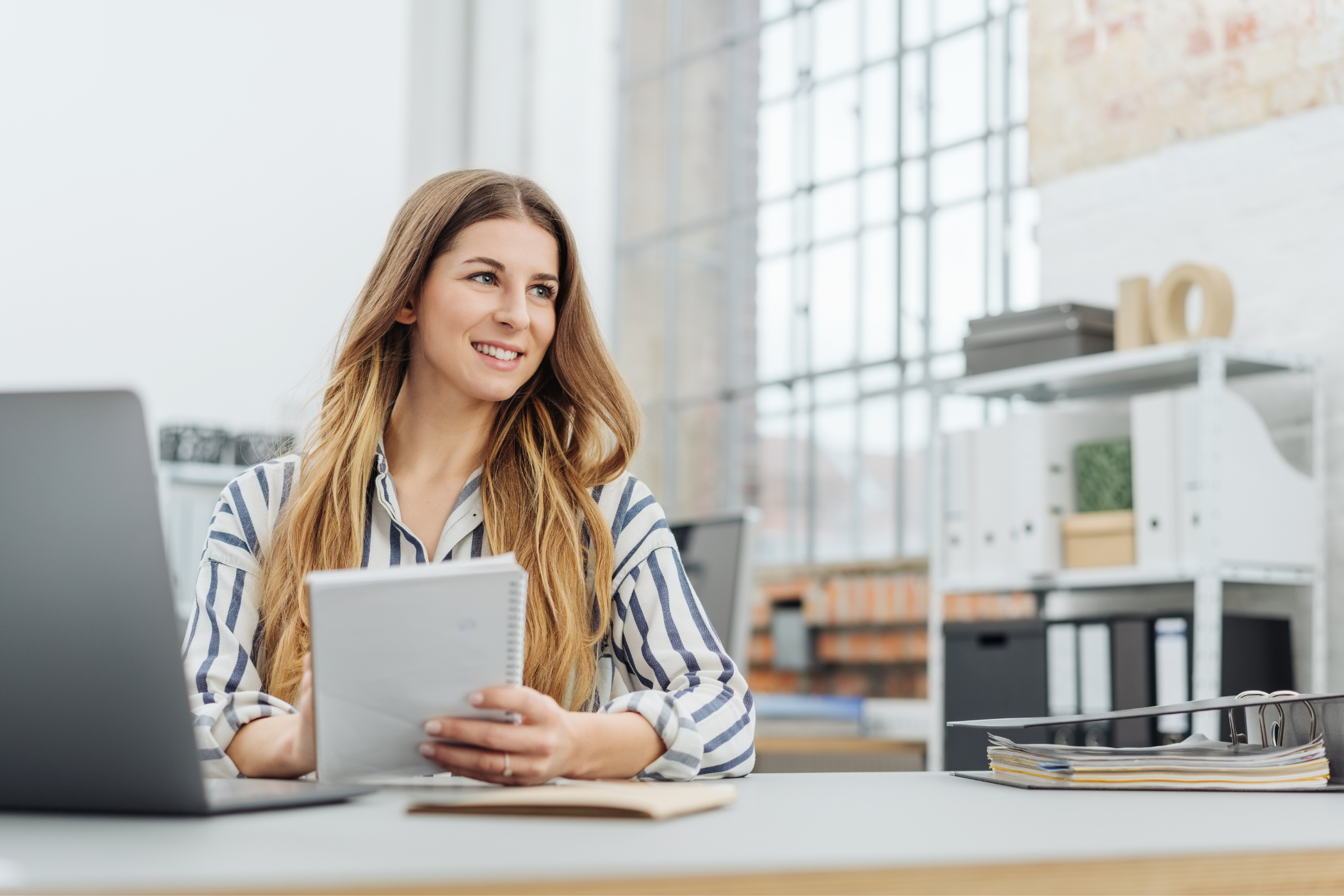 Smiling woman in a bright office holding a tablet at a desk with shelves and books in the background
