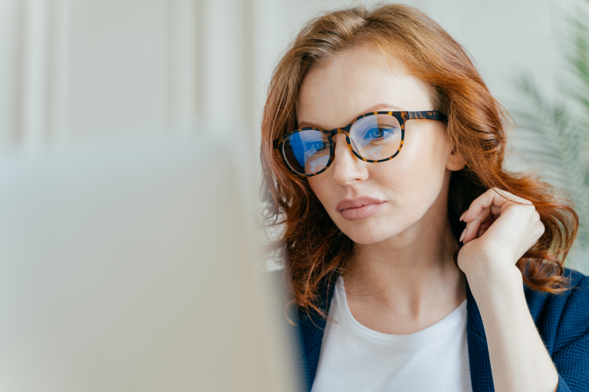 Woman with red hair and glasses working on a laptop, looking focused in a bright office