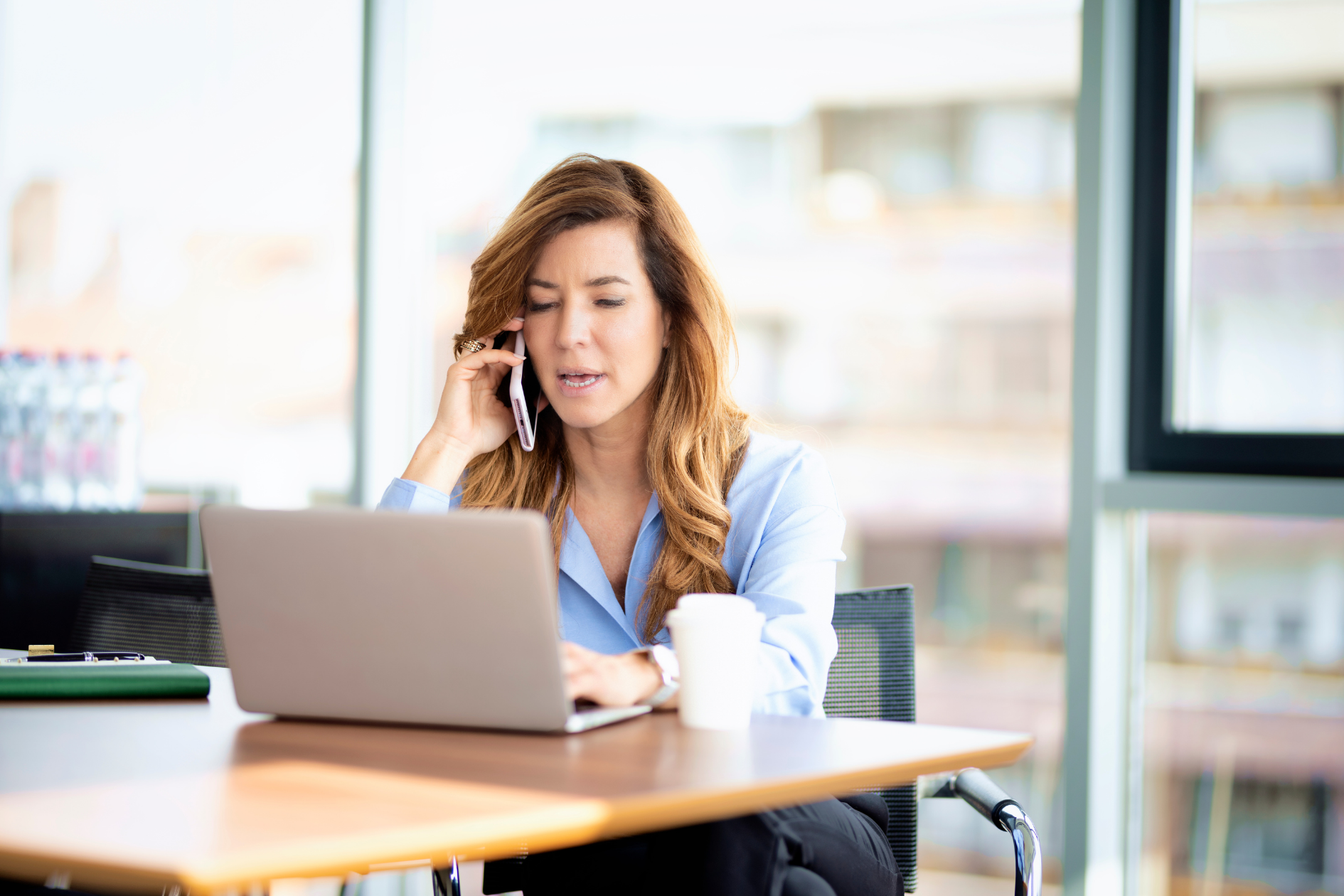 Woman at a laptop, looking stressed and talking on the phone in a bright office.
