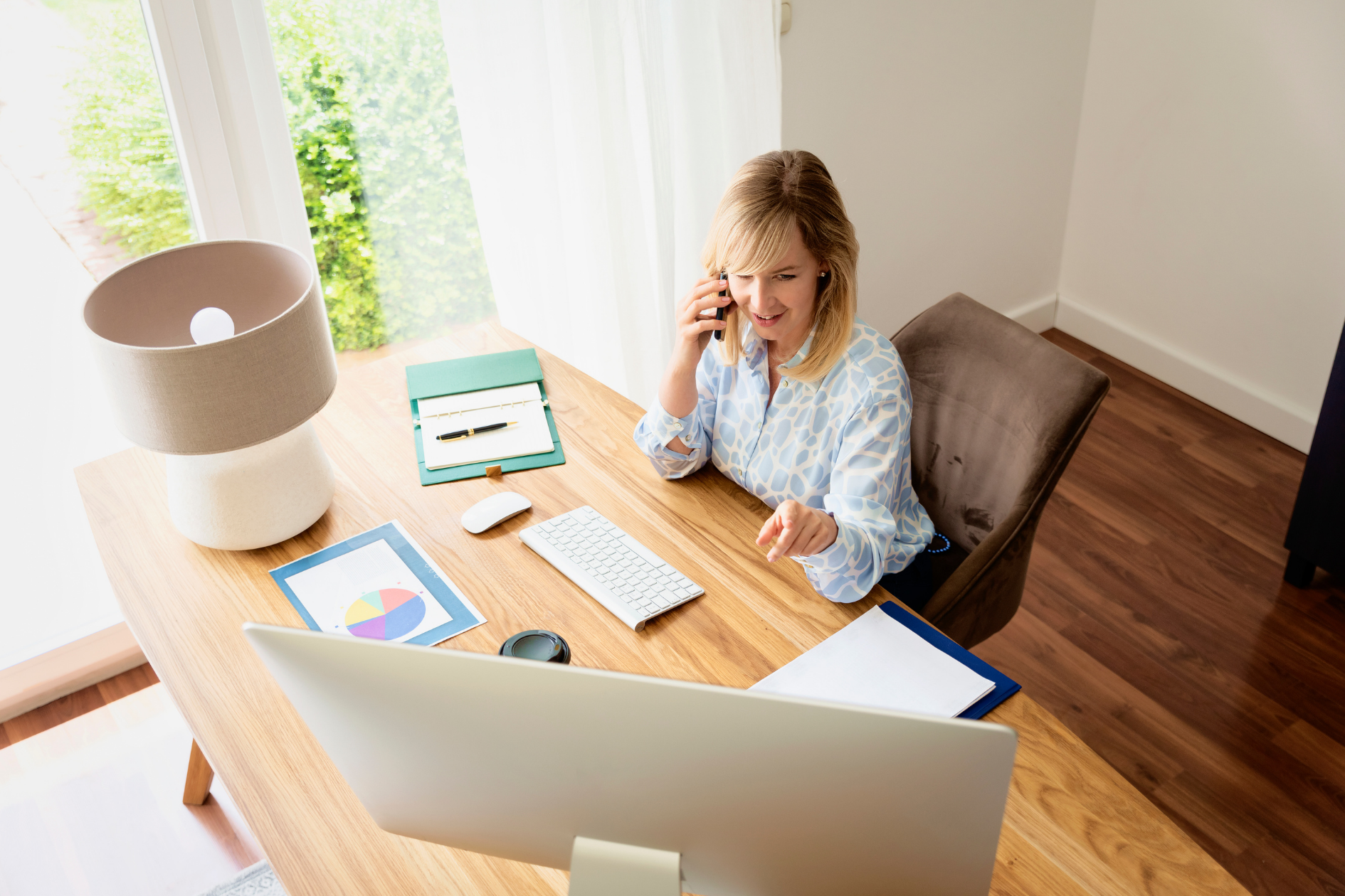 Person smiling at a desk in a bright home office, holding a phone and facing a computer monitor