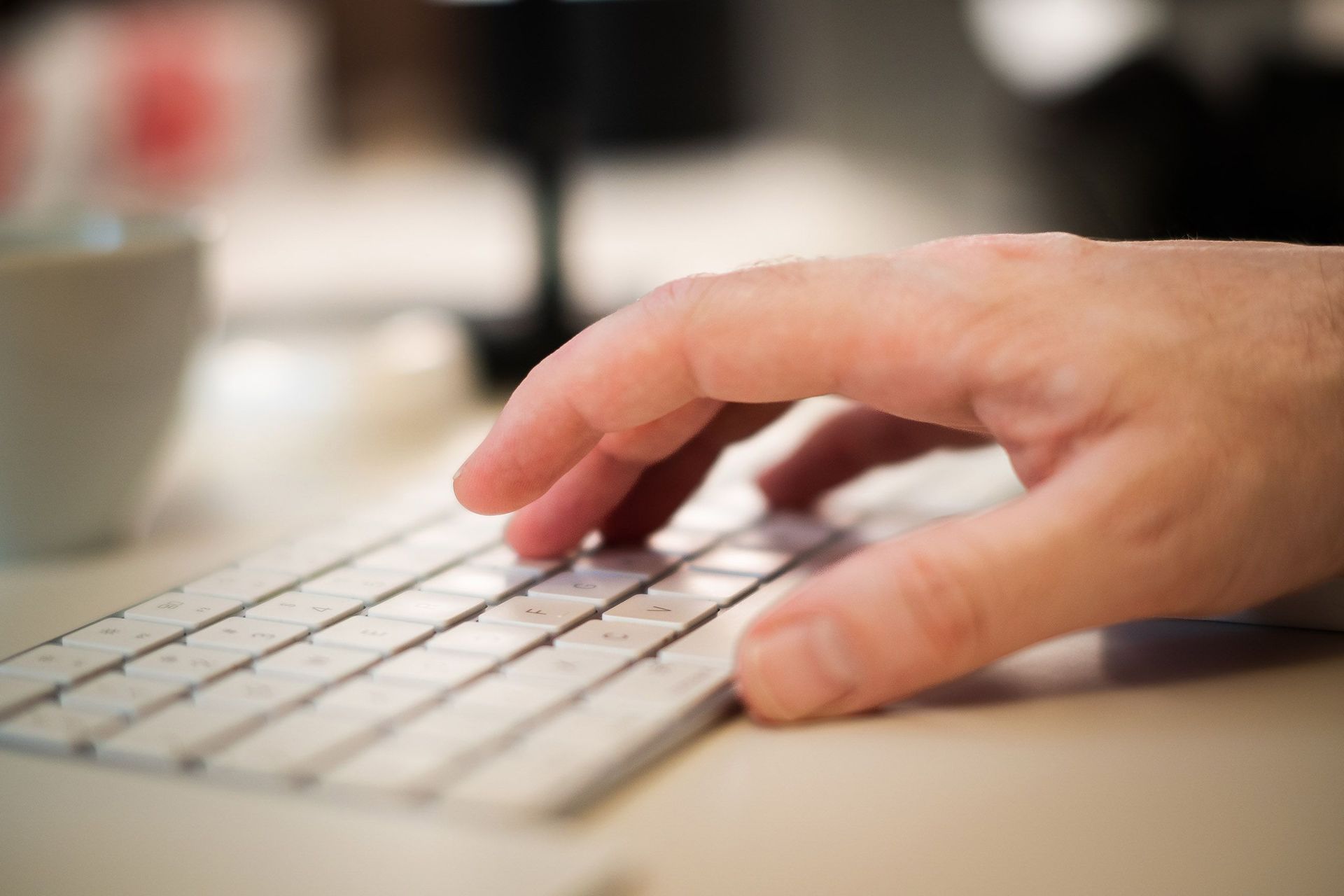 Hand typing on a white keyboard with a blurred coffee cup in the background