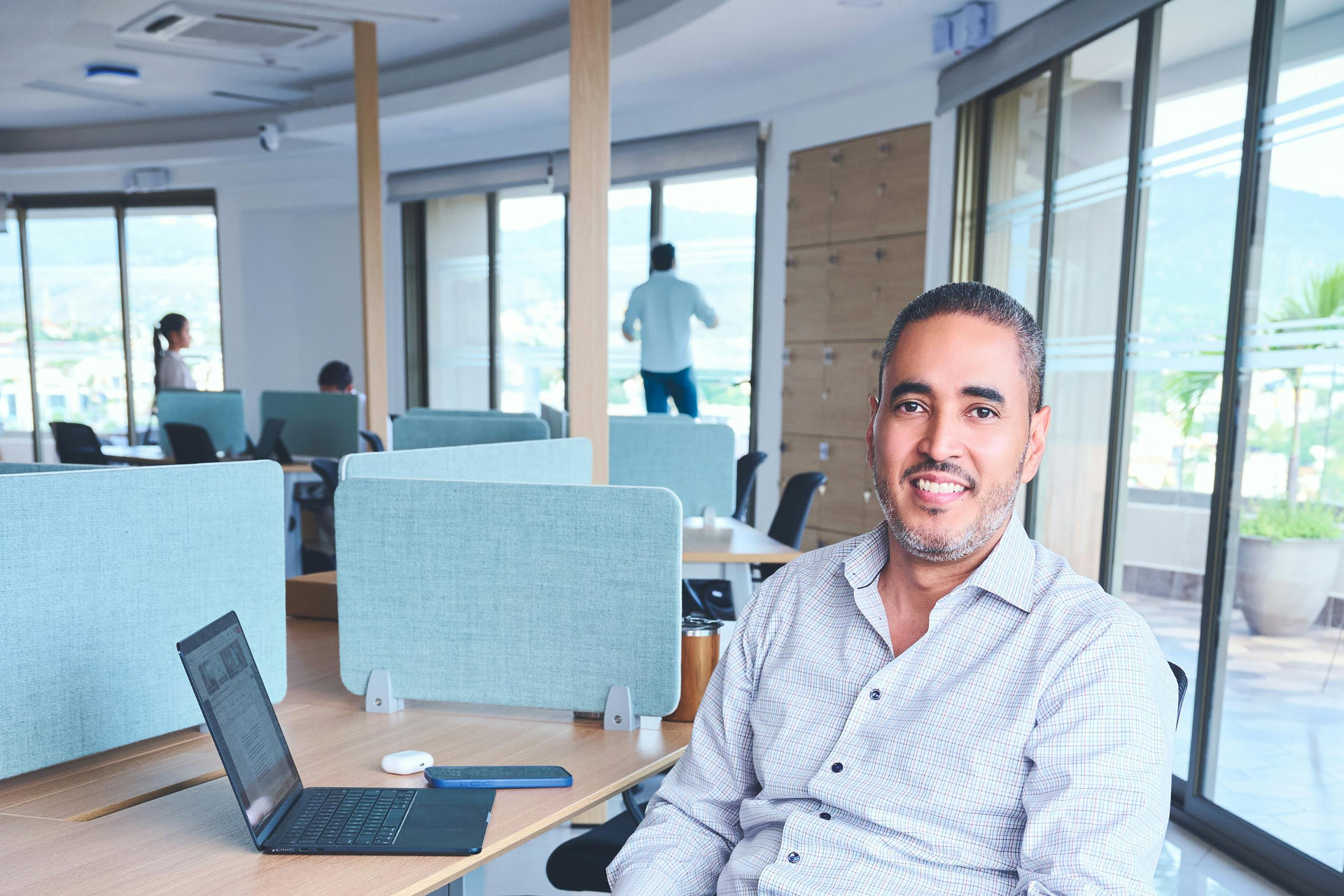 Man smiling at a desk in a bright office with a laptop and large windows