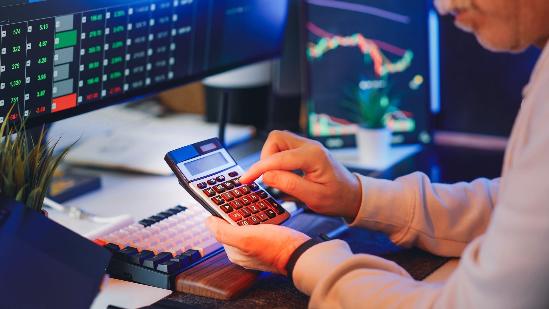 Hands using a calculator at a desk with financial charts on multiple monitors.