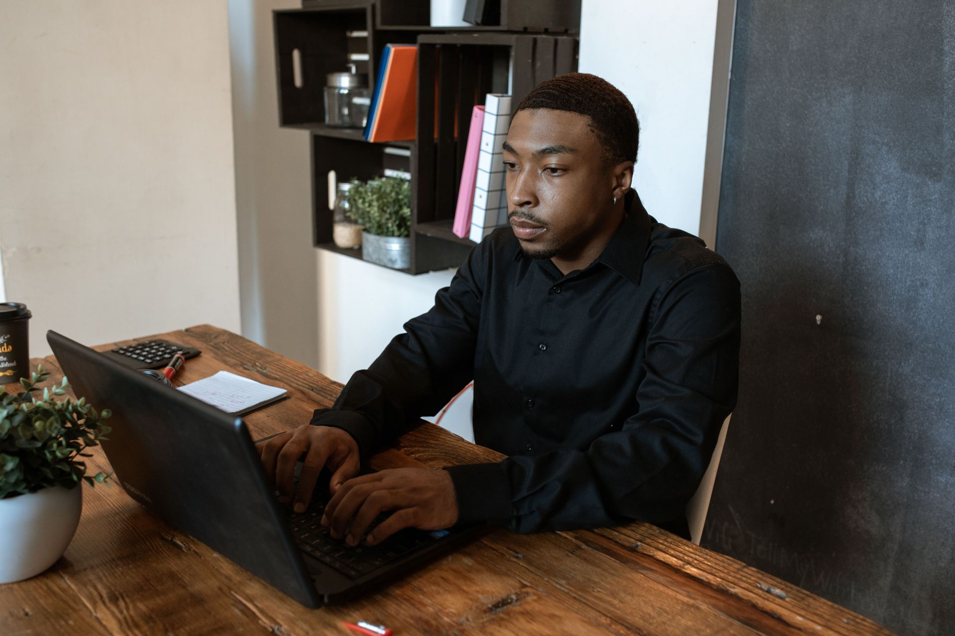Person typing on a laptop at a wooden desk in a modern office, with shelves and a plant nearby.