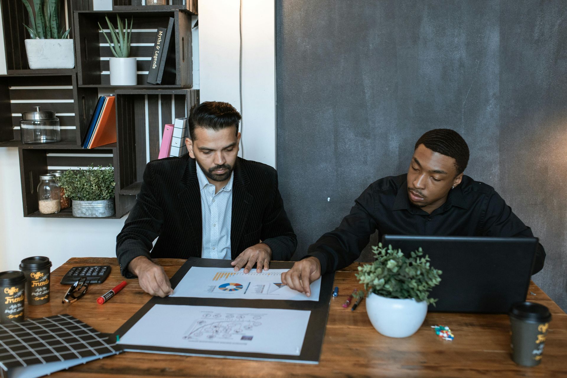Two men reviewing documents at a desk; one points to a graph. Wooden desk in a modern office.