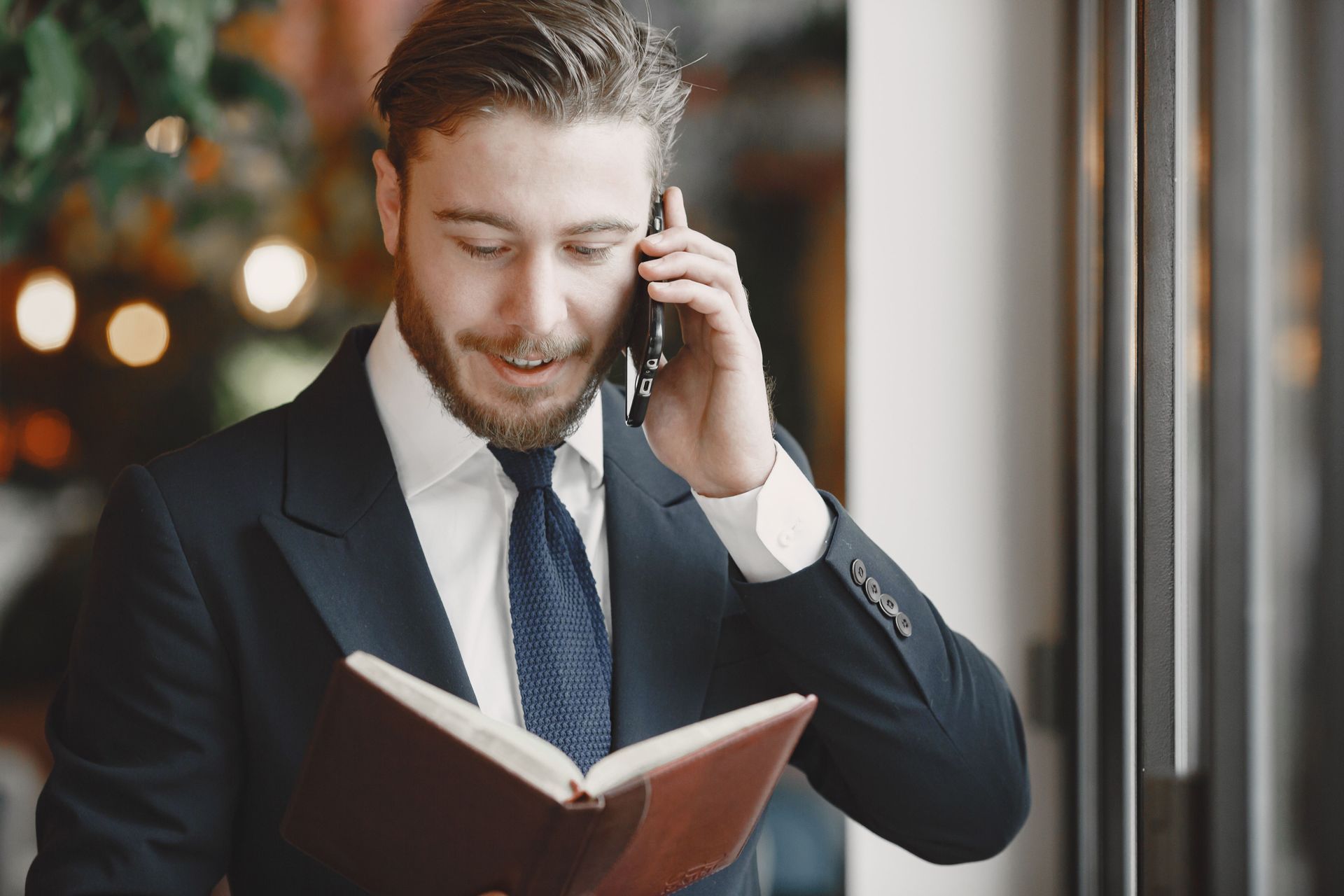 Businessman in a dark suit talking on a phone while reading a red notebook indoors