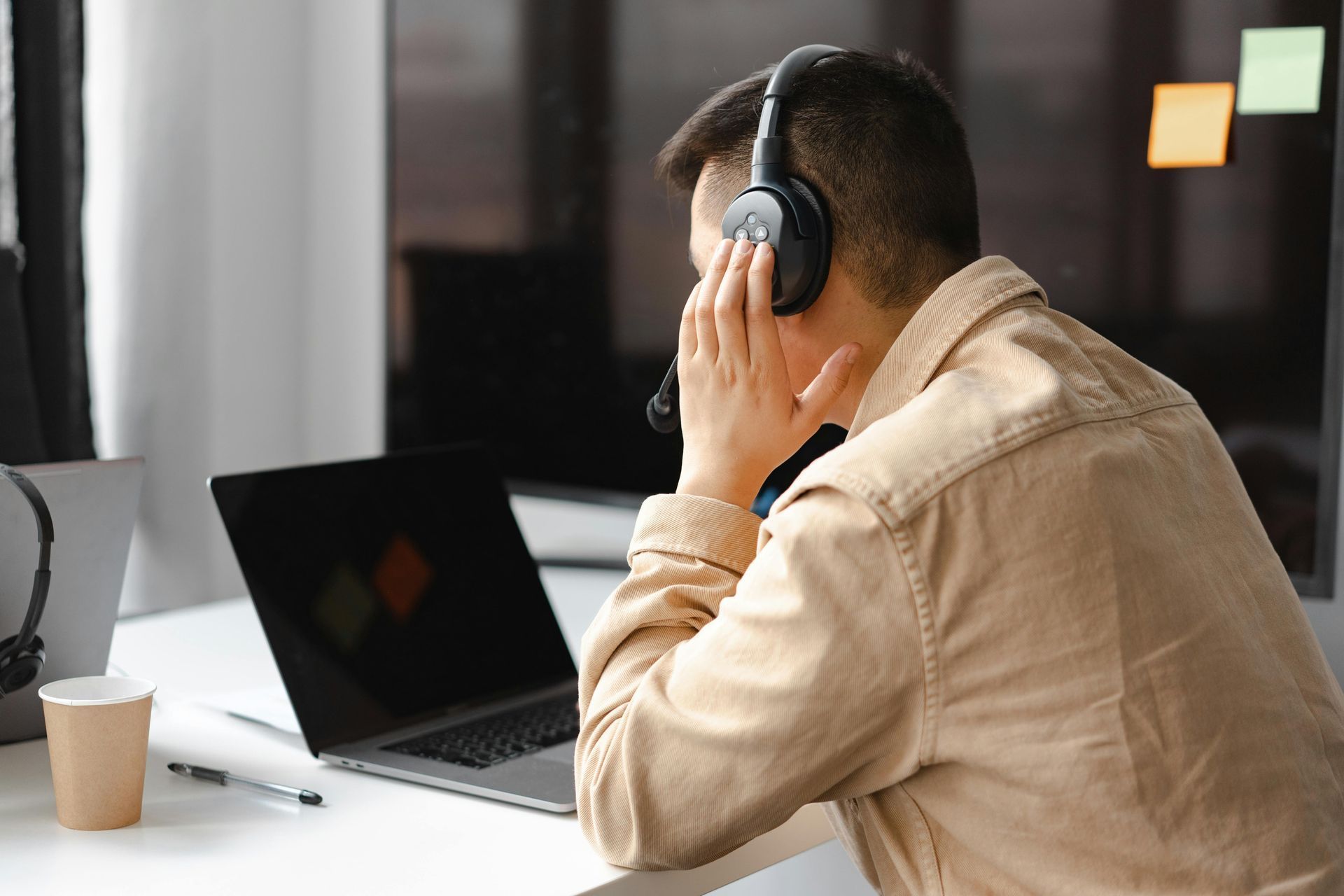 Person wearing headphones at a desk with a laptop, holding their head in a quiet office.