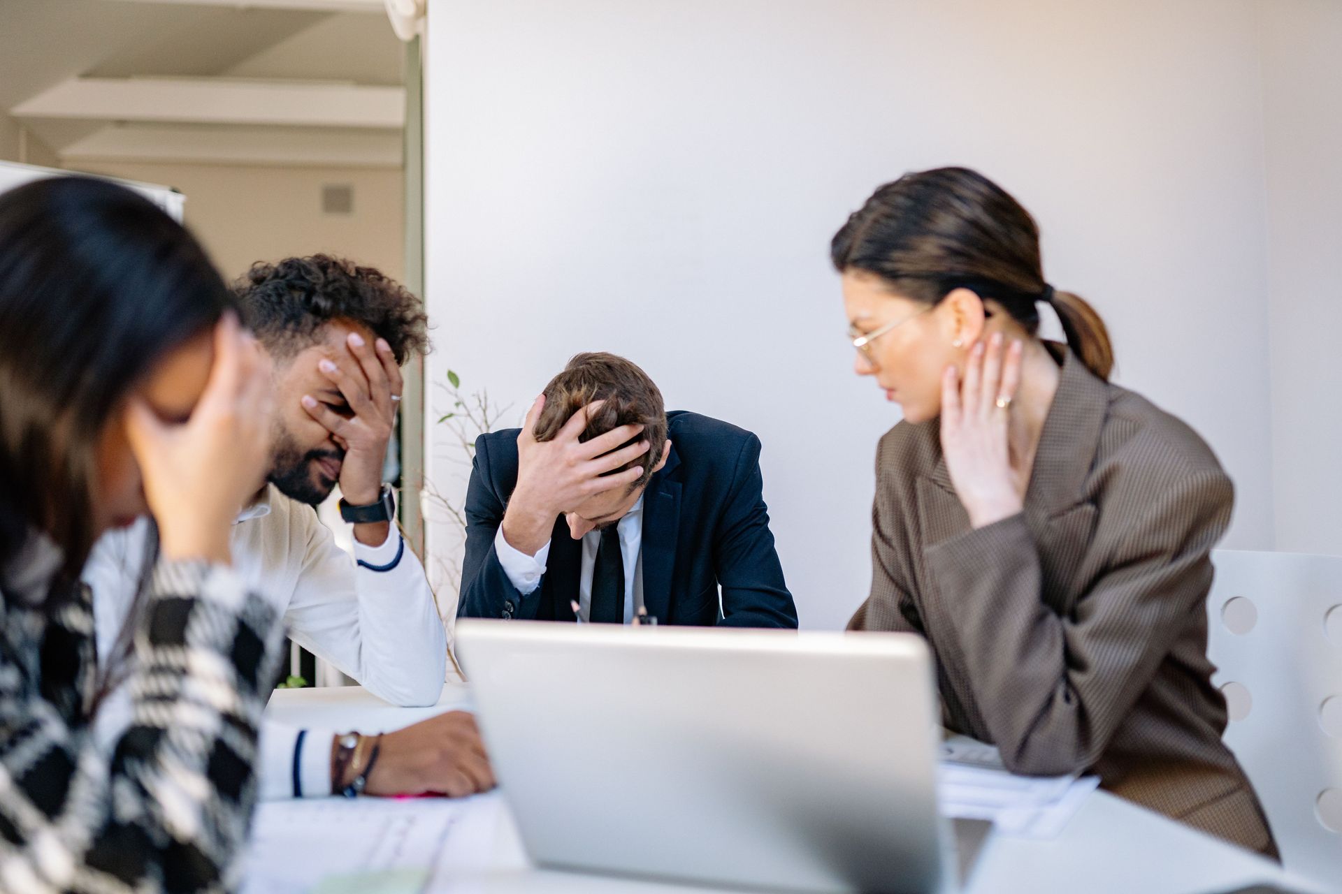 Stressed coworkers in a meeting room, two with heads in hands beside a laptop.