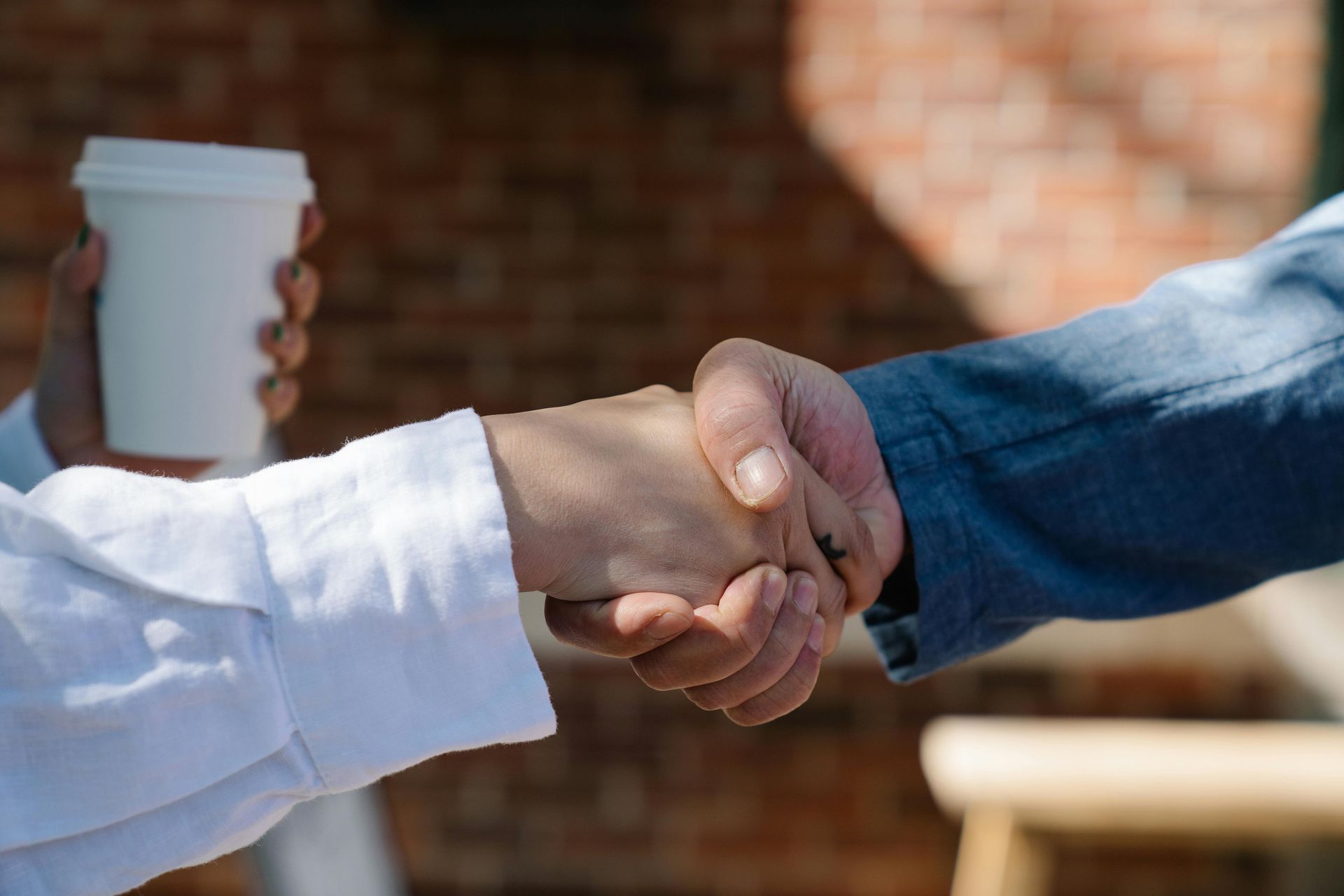 Two people shaking hands outdoors, one holding a white coffee cup in the background.