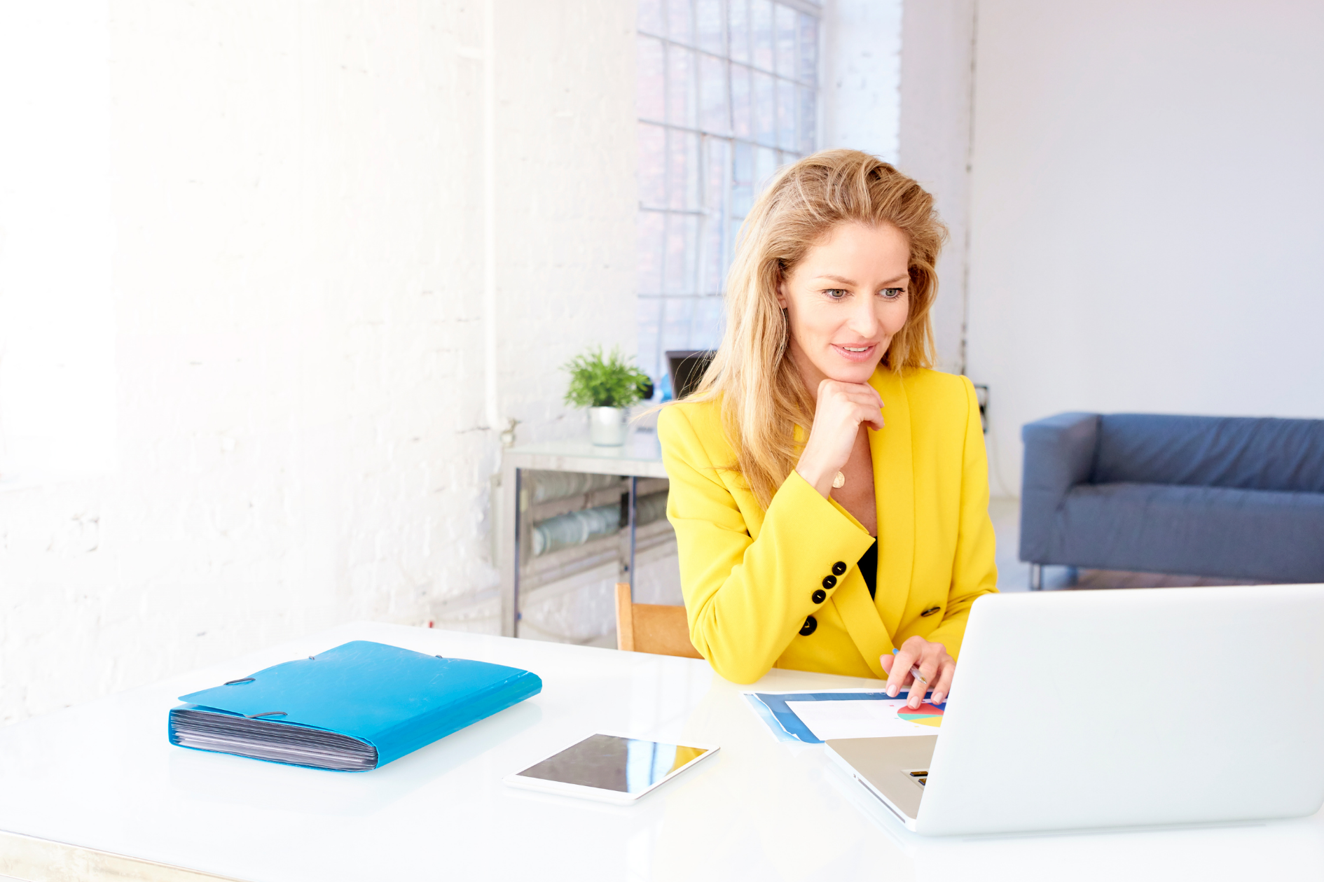 Woman in a yellow jacket working at a desk with a laptop in a bright office, smiling thoughtfully
