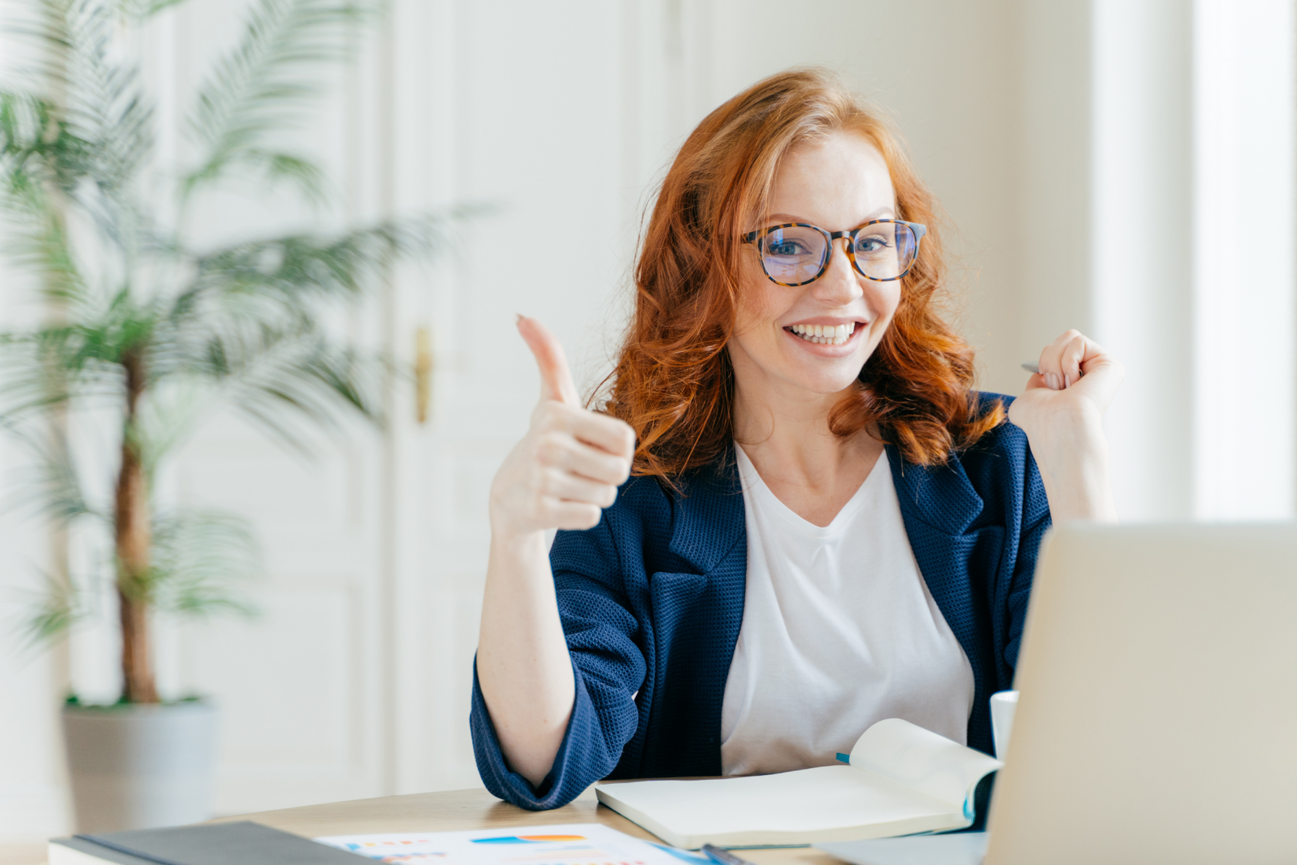 Smiling person at desk giving a thumbs-up beside an open laptop in a bright office