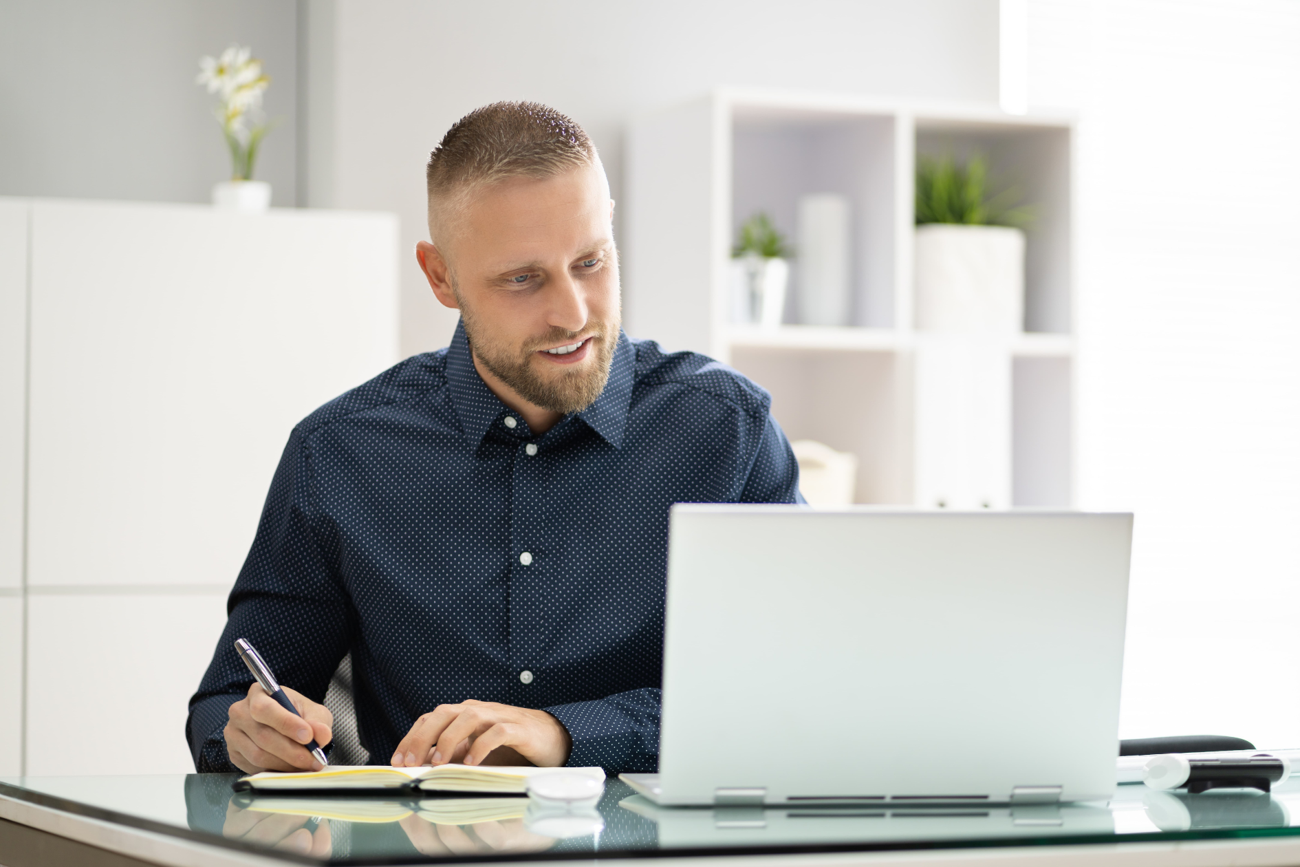 Man writing notes at a desk with a laptop in a bright office