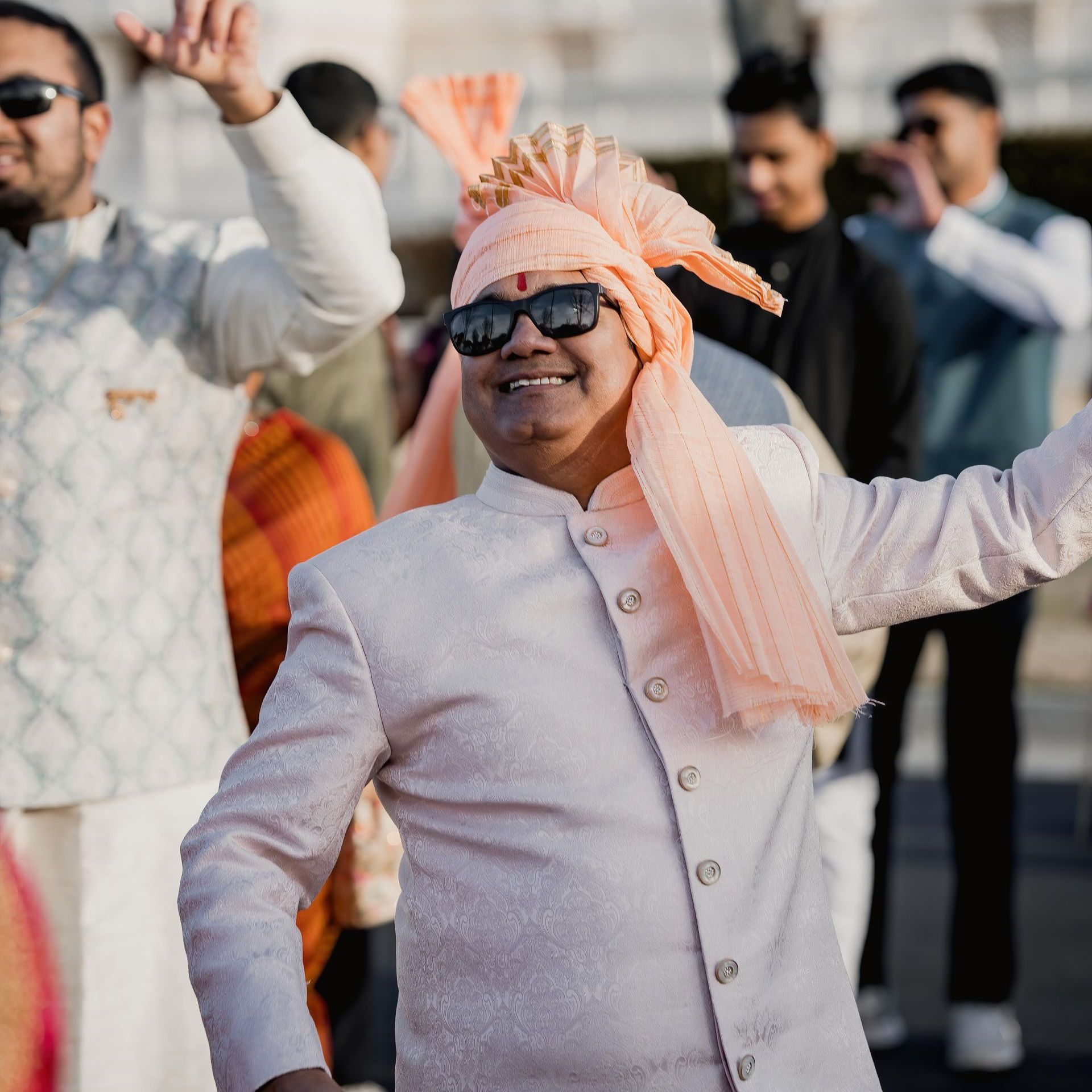 A man wearing sunglasses and a turban is dancing with his arms in the air.
