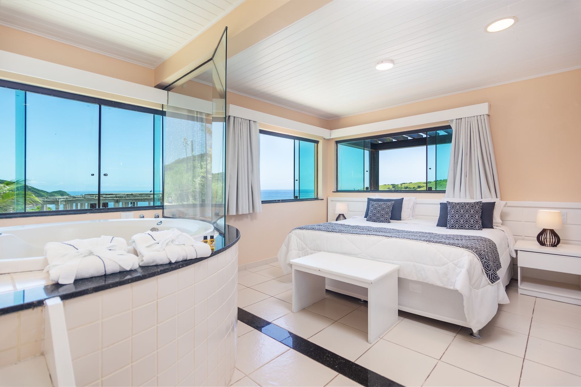 Quarto com jacuzzi, janelas amplas com vista para o mar, cama branca e decoração moderna.
