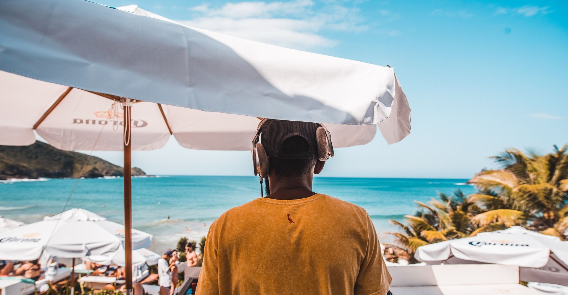 DJ em uma festa na praia, com vista para o mar, guarda-sóis brancos e céu limpo.