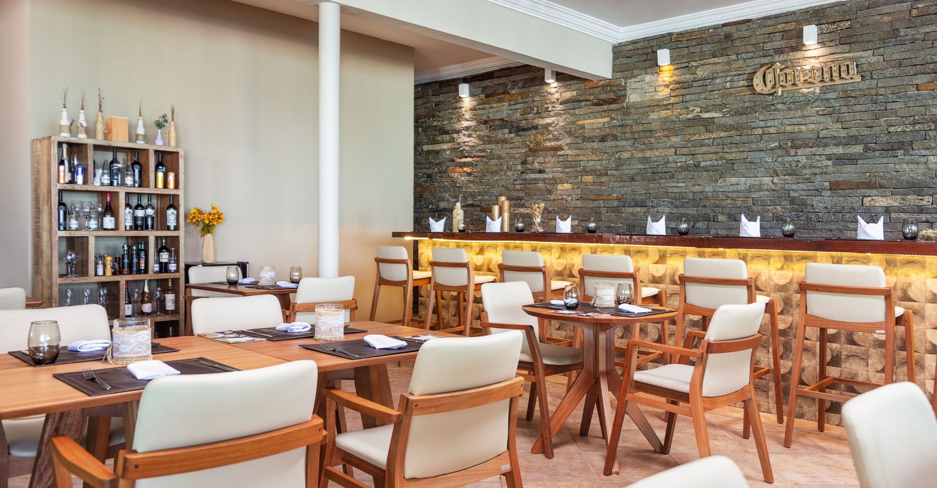 Interior de restaurante com mesas e cadeiras de madeira, balcão com banquetas e adega. Parede com detalhe em pedra.