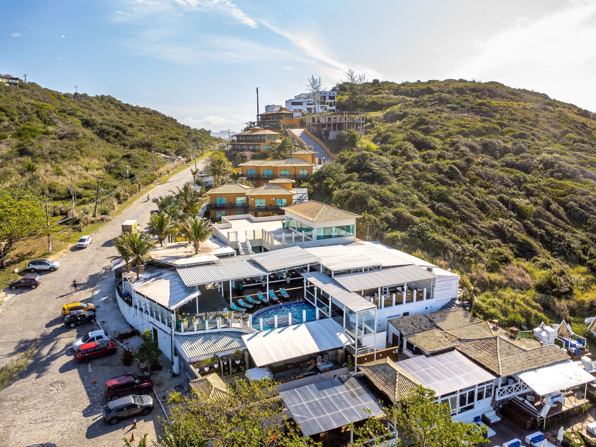 Restaurante branco com piscina em uma encosta, com outros edifícios e uma estrada visíveis. Dia ensolarado.