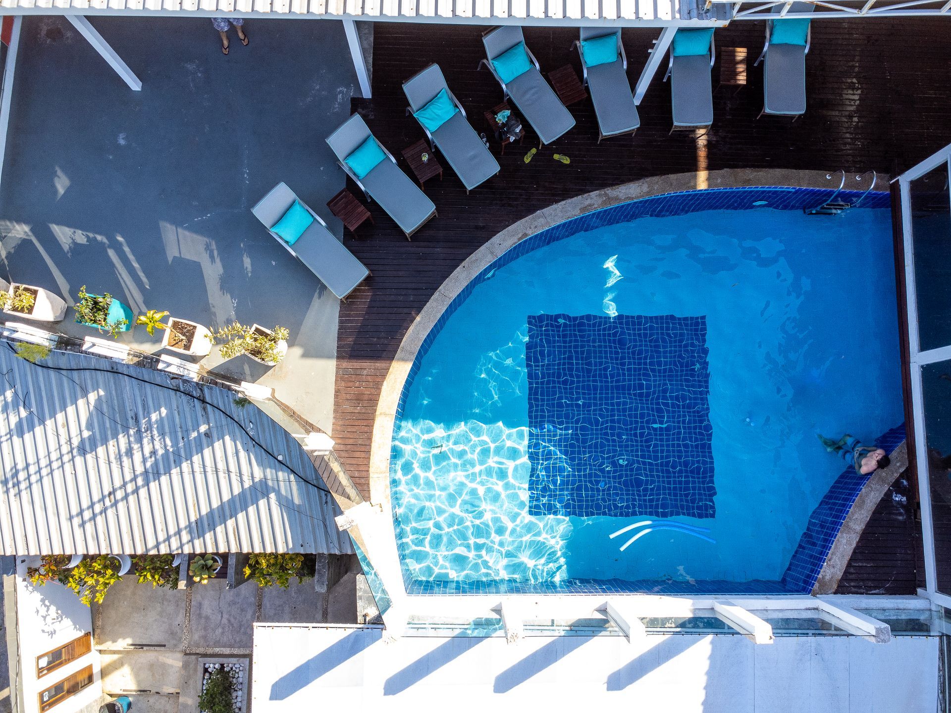 Vista aérea de uma piscina com espreguiçadeiras e deck sombreado. Tons de azul e marrom.