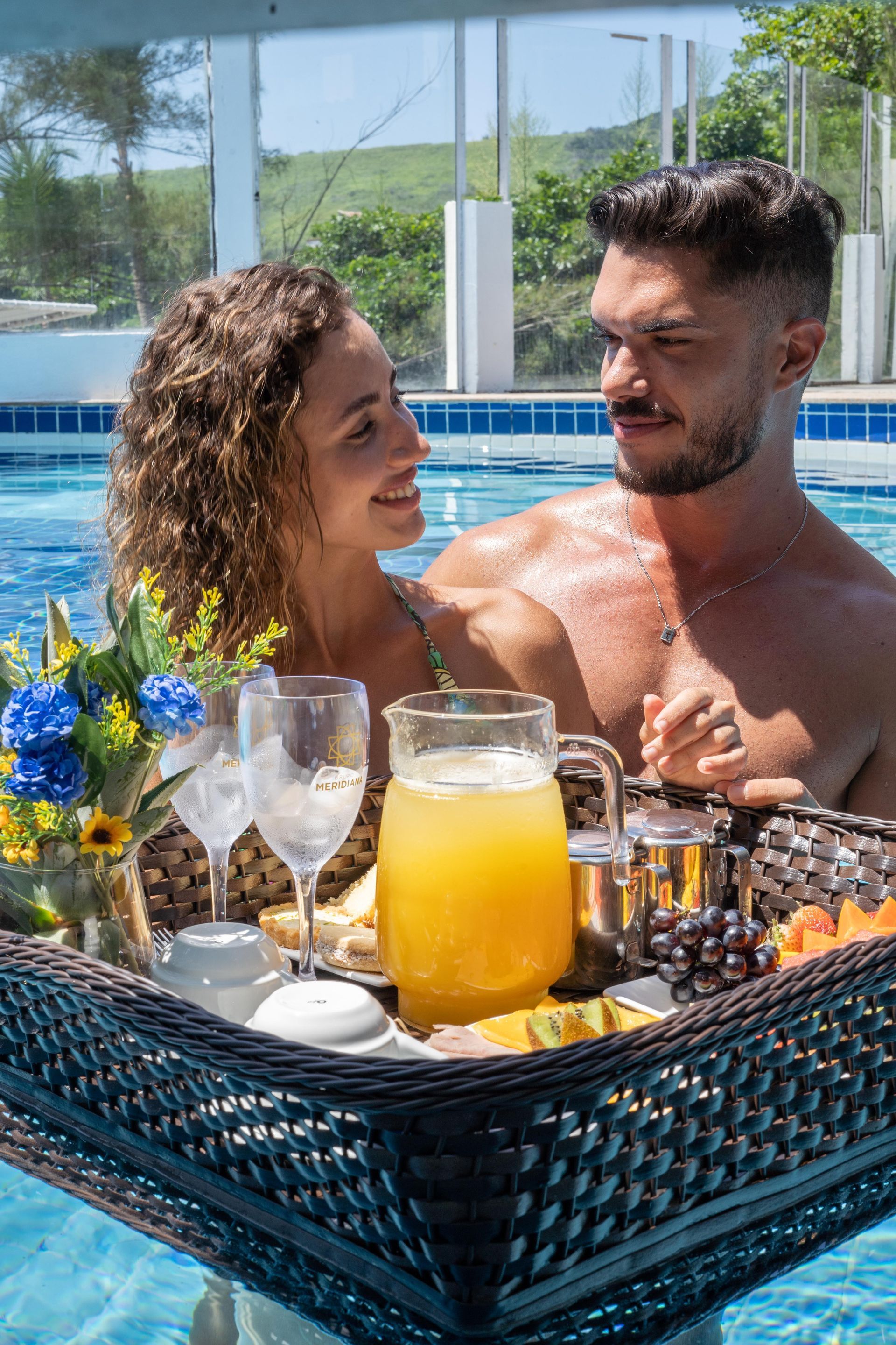 Casal relaxando na piscina, tomando café da manhã em uma bandeja flutuante, ensolarados e sorridentes.