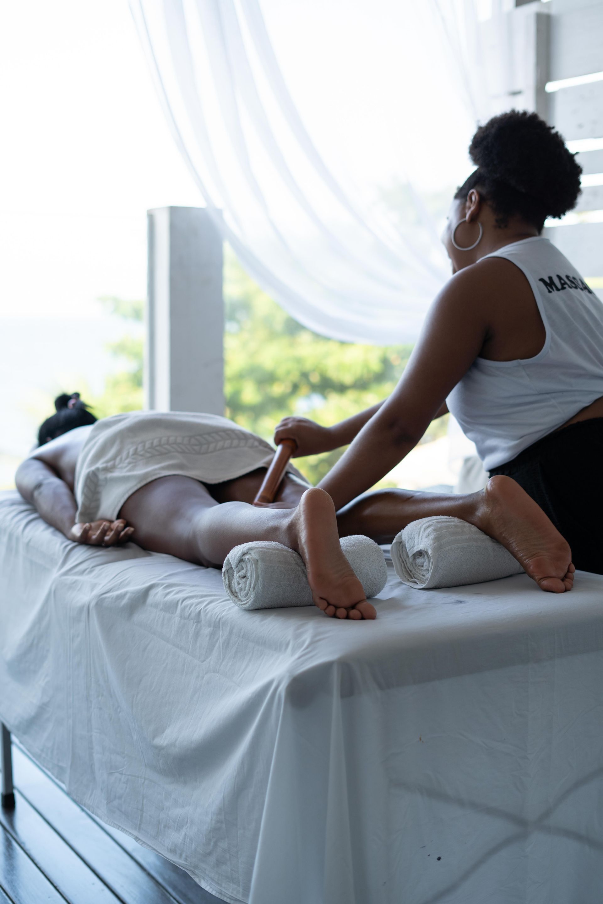 Massagista realizando uma massagem nas pernas em uma maca. Ambiente externo amplo e iluminado.