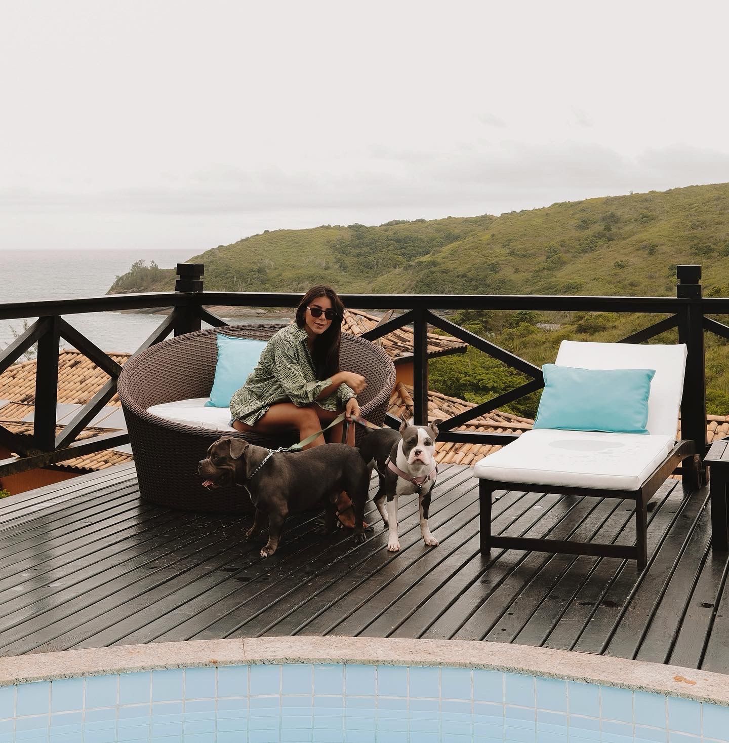 Mulher com dois cachorros em um deck com vista para o litoral, perto de uma piscina. Céu nublado.