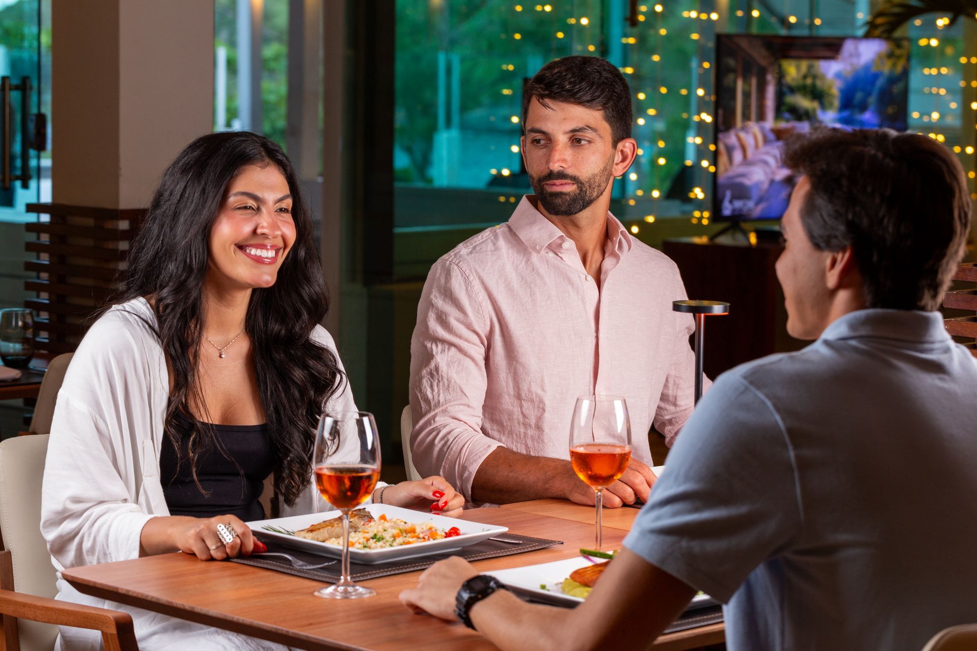 Três pessoas sentadas à mesa de um restaurante, jantando e conversando; a mulher sorrindo, o homem observando.