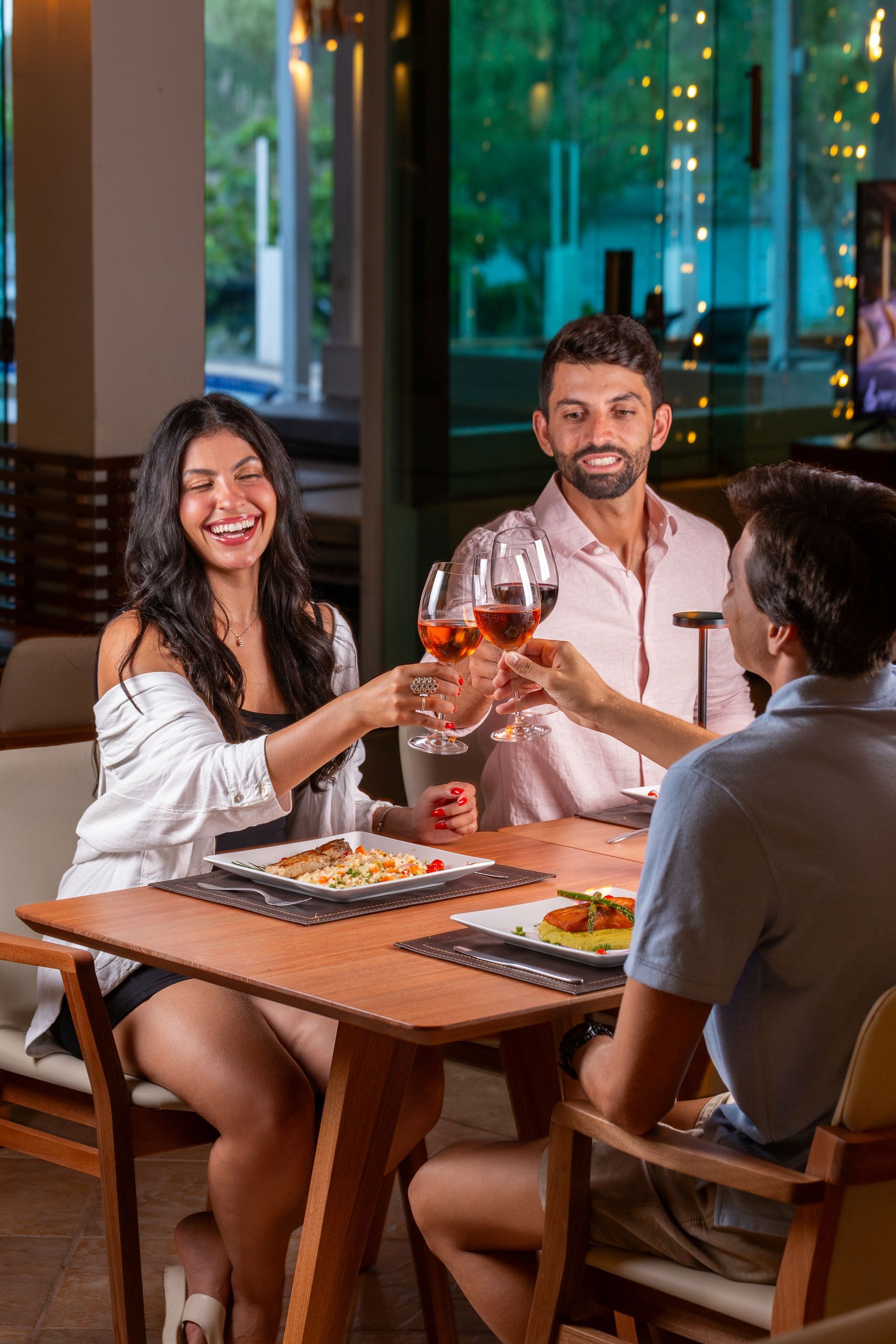 Três pessoas brindando com taças de vinho à mesa. Rostos sorridentes, pratos de comida, mesa de madeira.