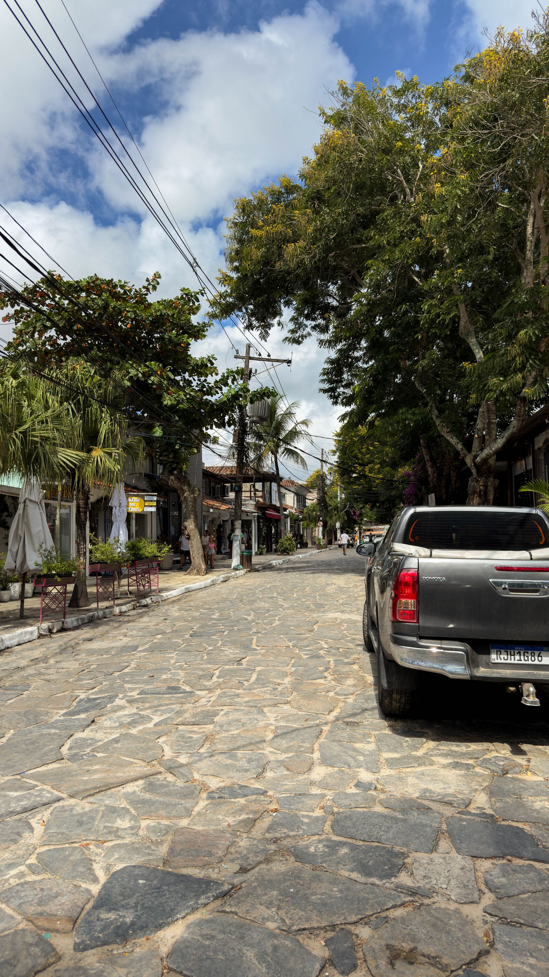 Rua de paralelepípedos ladeada por prédios e árvores, um caminhão estacionado à direita sob um céu parcialmente nublado.