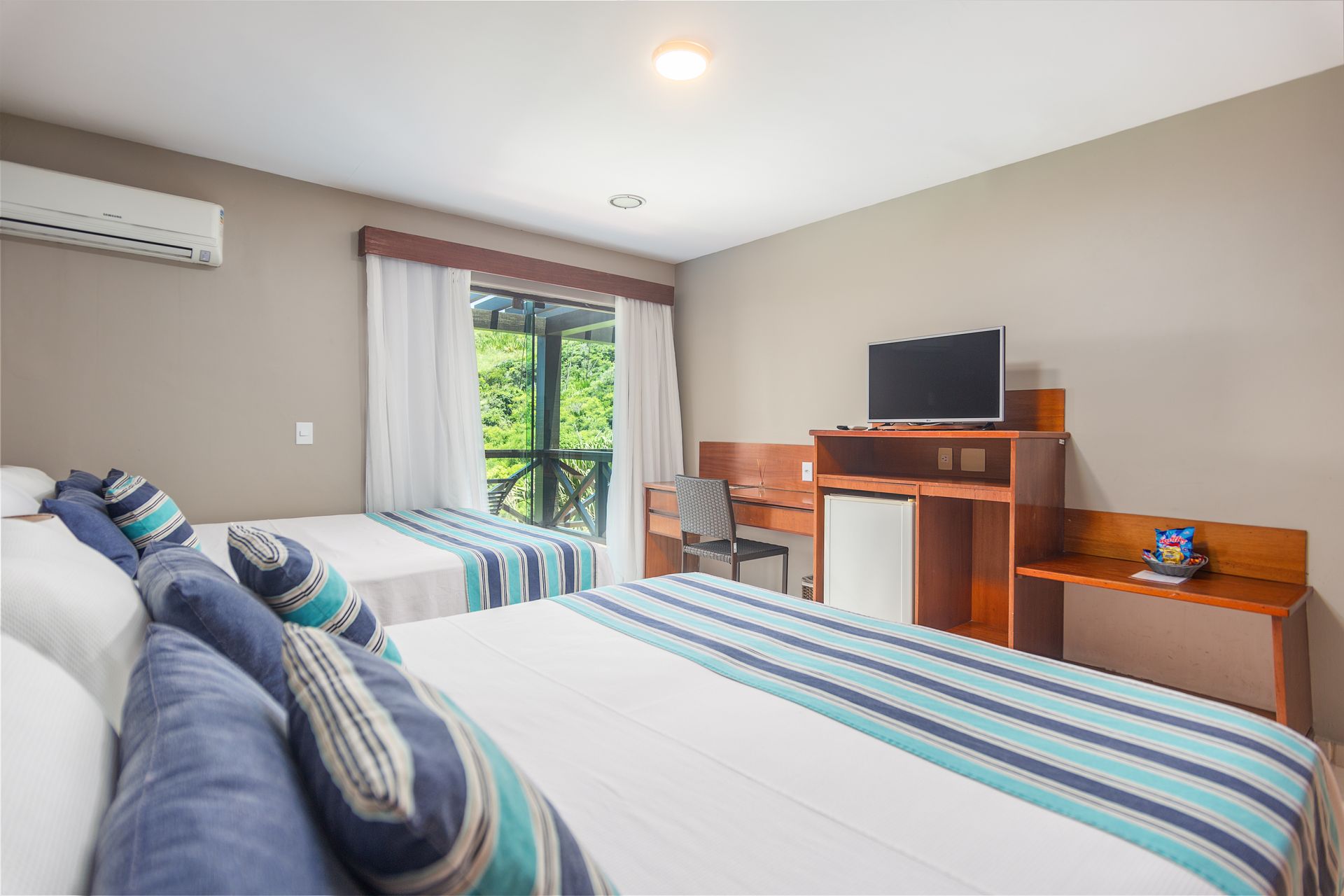 Quarto de hotel com duas camas, escrivaninha, TV e vista para a vegetação através da janela.