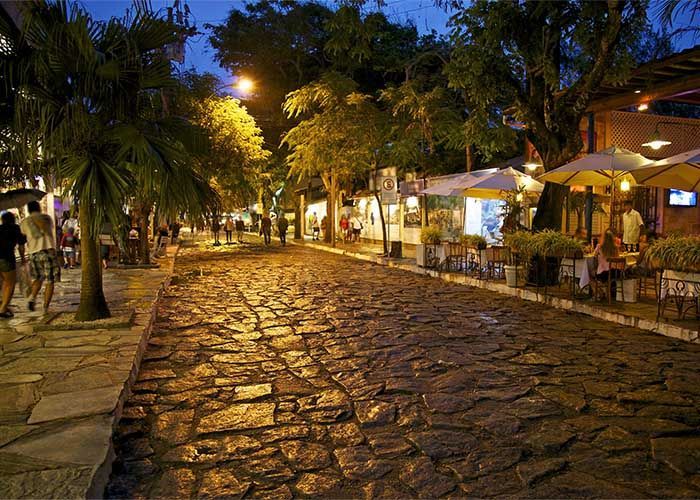 Rua de paralelepípedos à noite, repleta de restaurantes e palmeiras, iluminada por postes de luz.