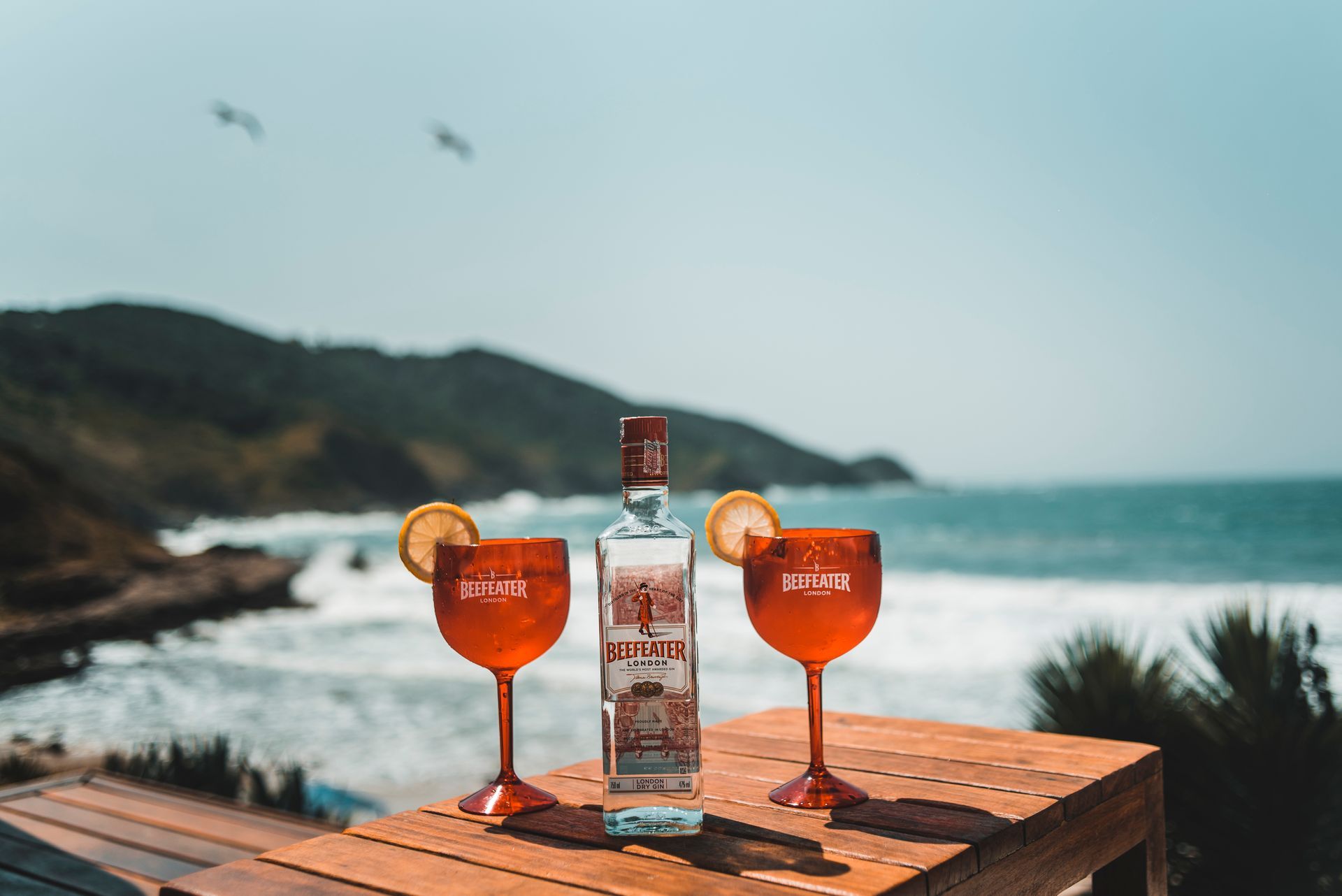Garrafa de bebida alcoólica e dois copos vermelhos com rodelas de limão sobre uma mesa de madeira, tendo como pano de fundo a praia e o oceano.
