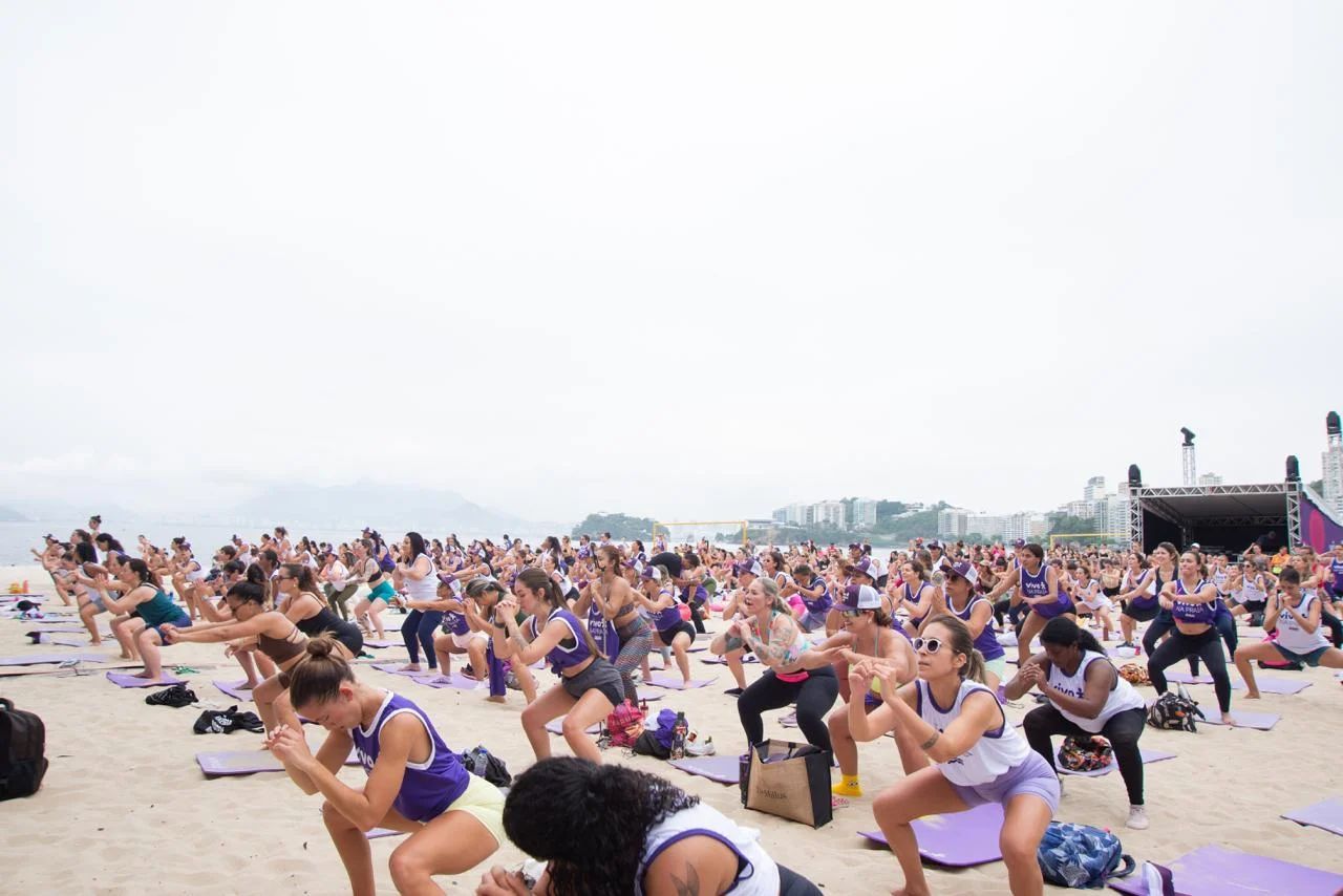 Large group of people exercising on a beach, performing squats on yoga mats near a stage.