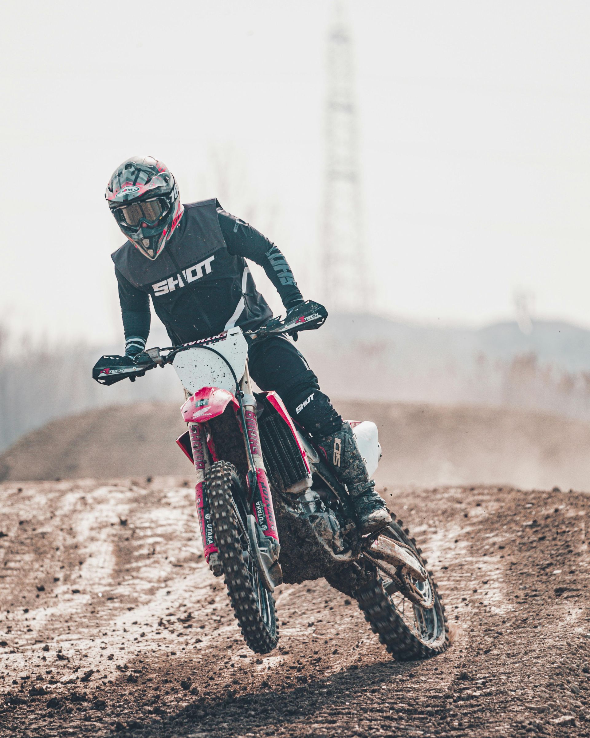 Piloto de motocross apoiado em uma pista de terra. Moto vermelha e branca, usando equipamento preto. Céu nublado ao fundo.