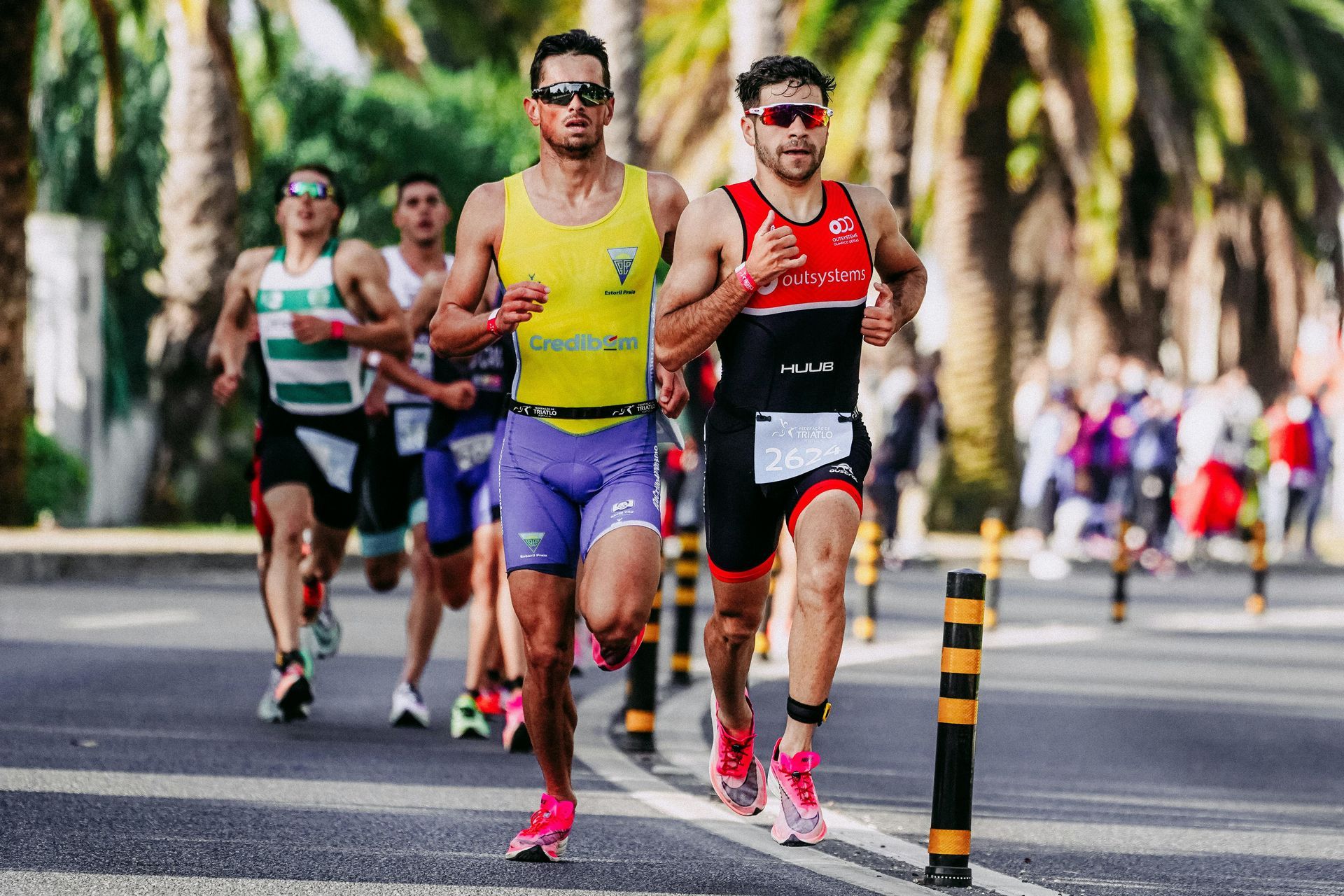 Several runners in athletic gear race down a paved road lined with palm trees.