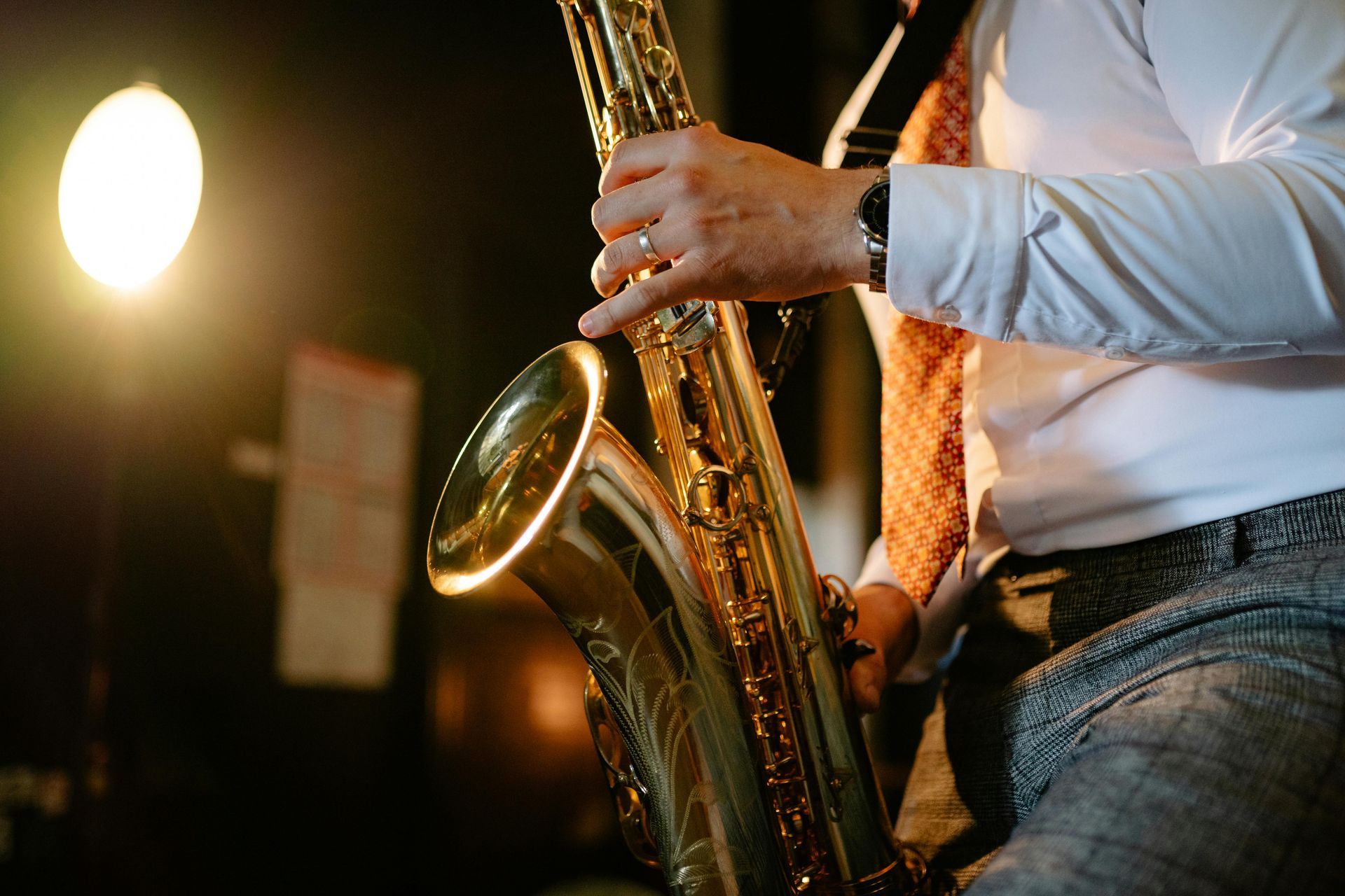 Pessoa tocando um saxofone dourado, iluminada por um holofote, vestindo camisa e gravata brancas.