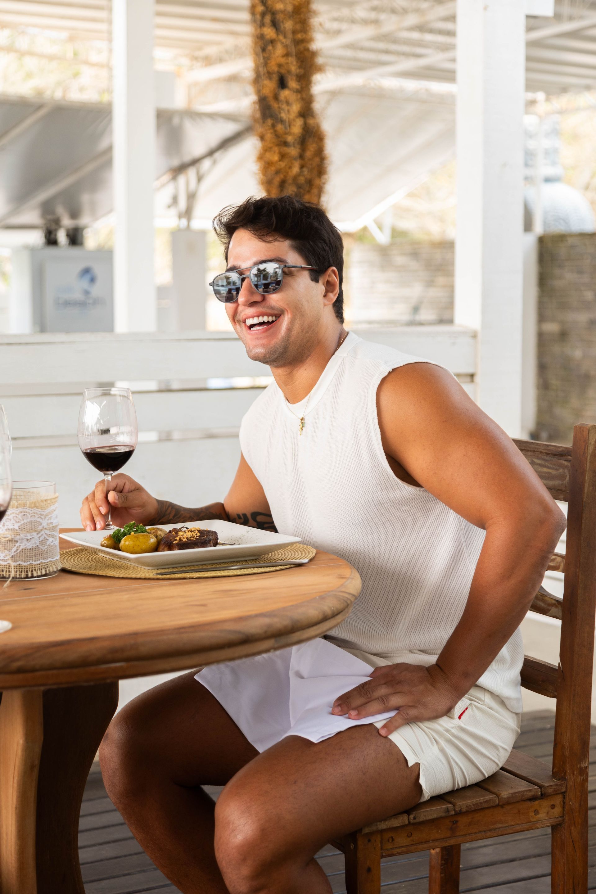 Homem sorrindo, jantando ao ar livre em uma mesa redonda, segurando uma taça de vinho. Usando óculos de sol