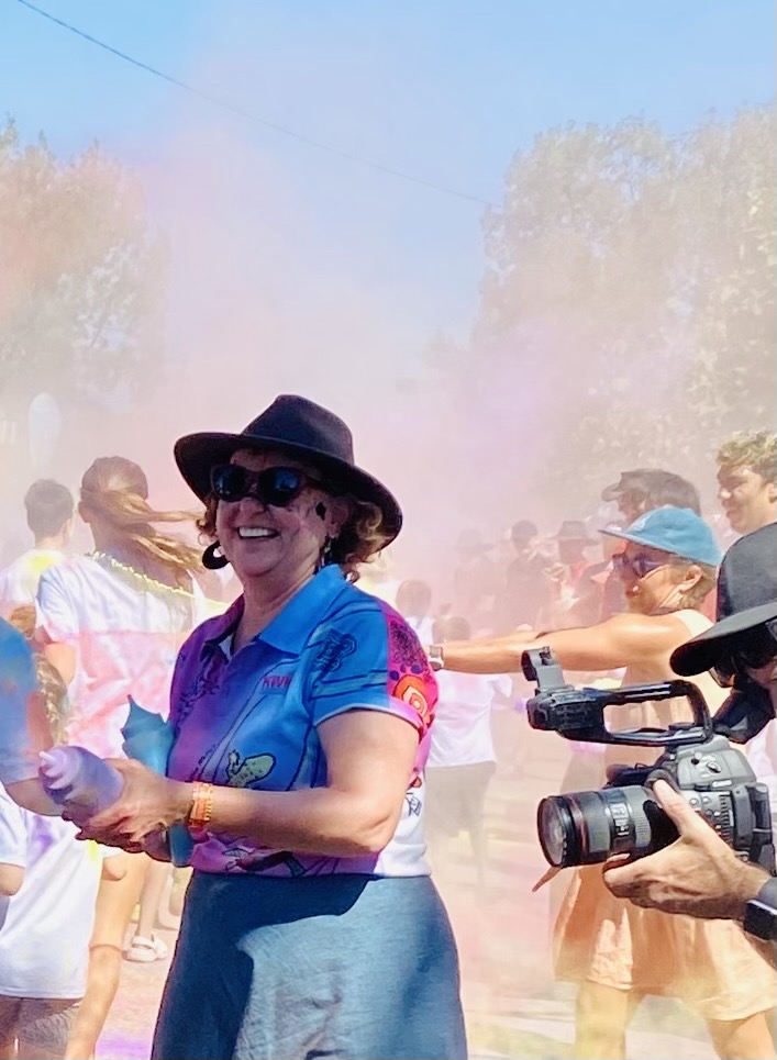 Woman in hat covered in colored powder at an outdoor event, smiling. Other people visible, some filming.