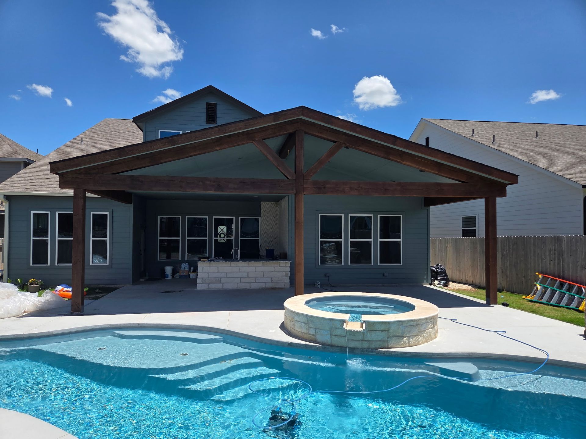 Backyard with pool, spa, and covered patio. Blue house with a brown roof, sunny day.