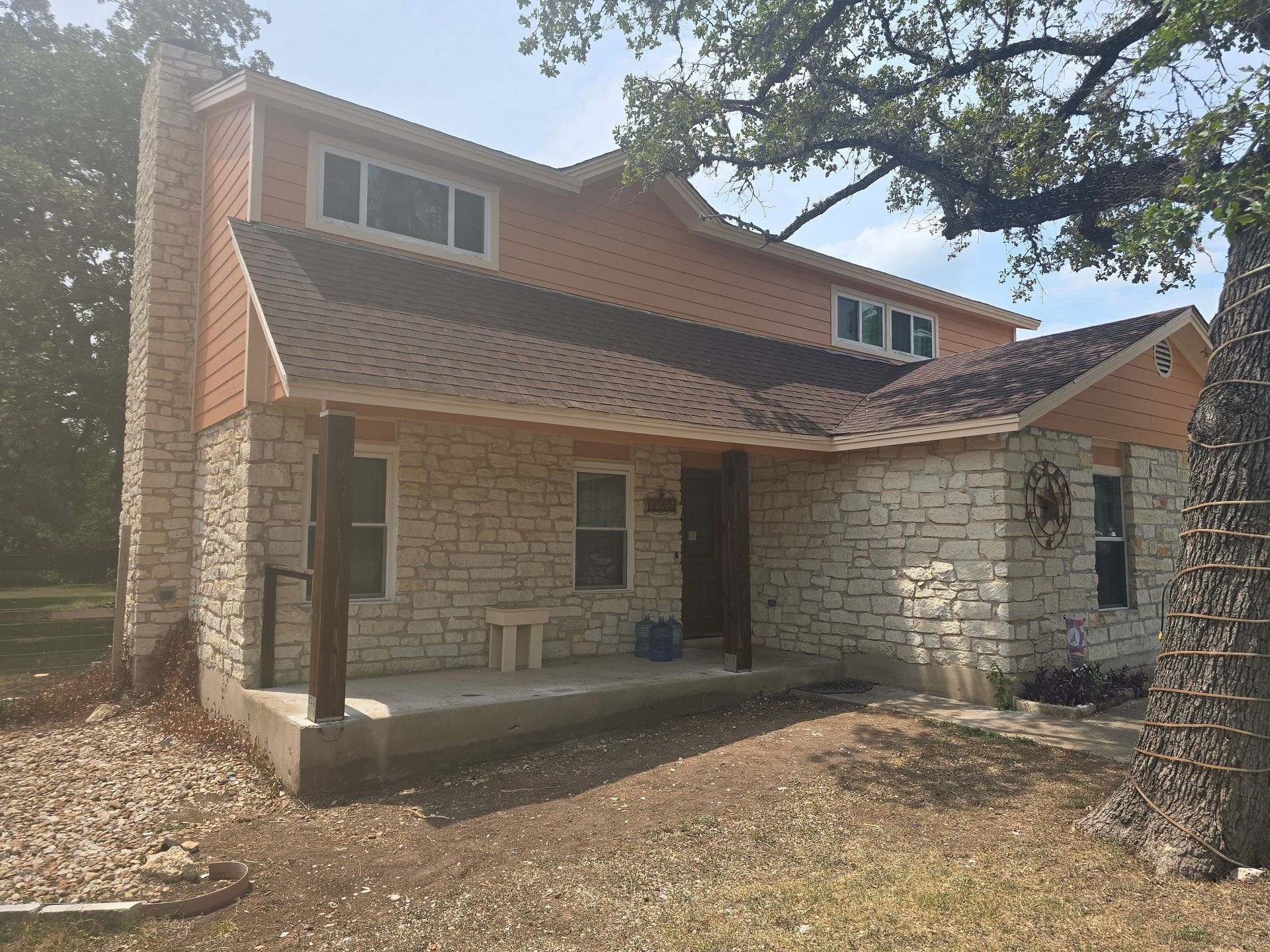 Two-story house with stone facade, tan siding, and dark brown roof; porch, chimney, and large tree.