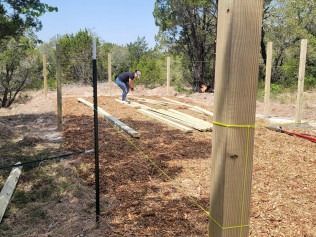 Person building a fence in a wooded area; brown wood posts, wood chips, bright sun.
