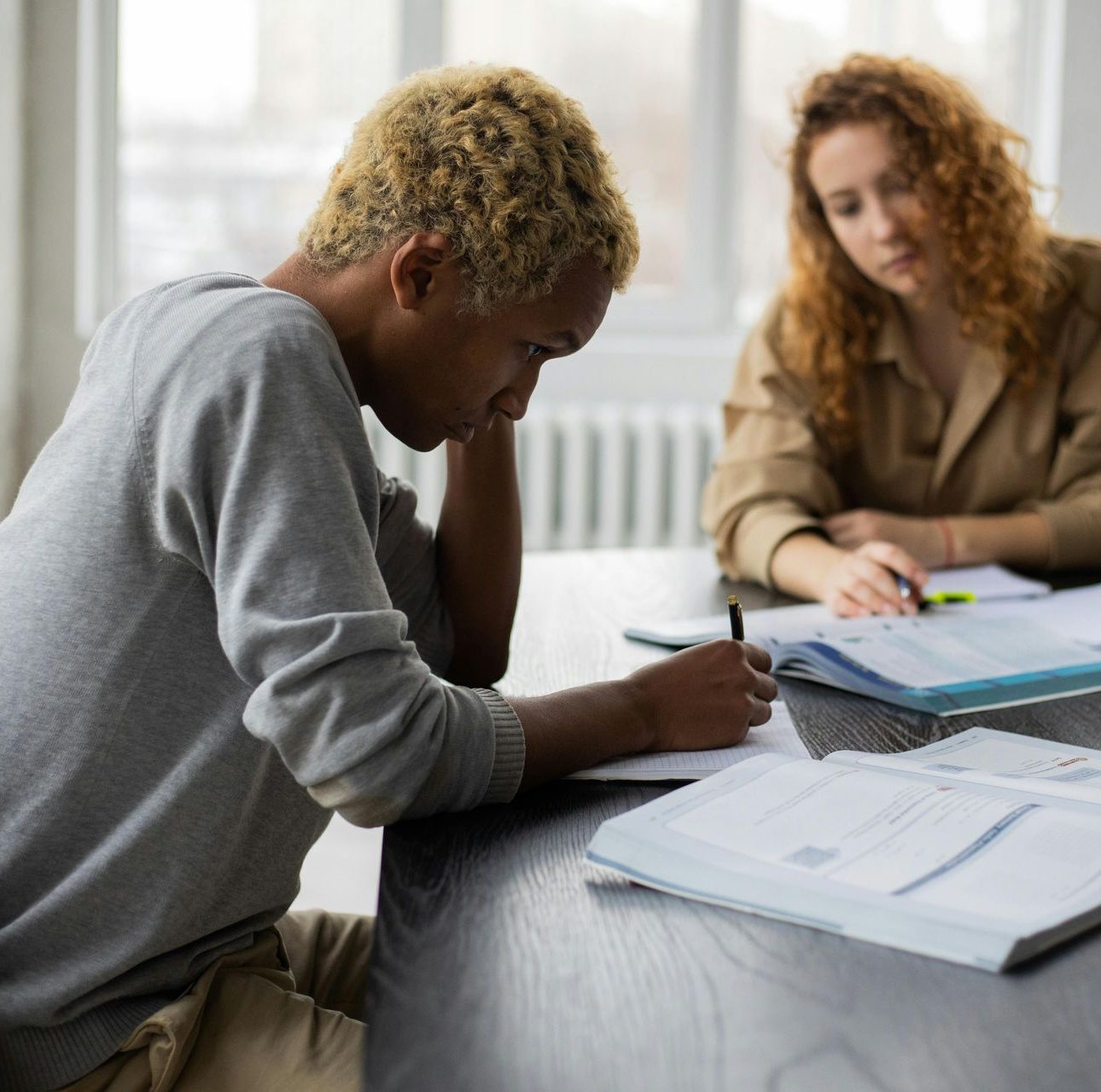 Student writing at a table while a teacher reviews papers beside them in a bright classroom