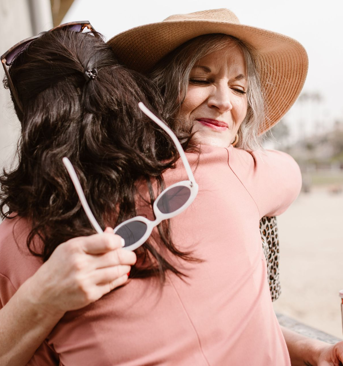 Two people hugging outdoors, one wearing a wide-brim hat and smiling while holding sunglasses.