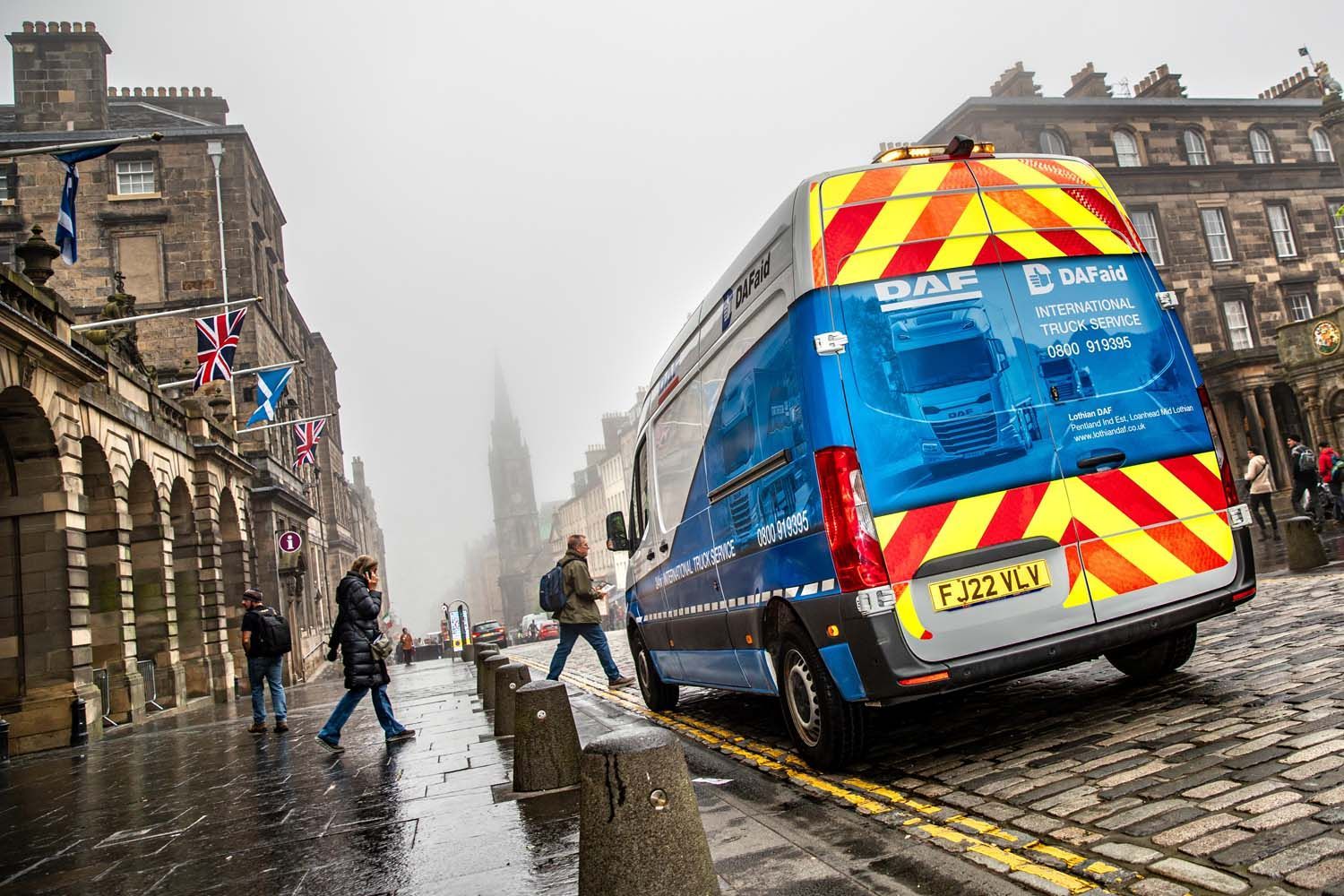 Transport photography of van on a wet cobblestone street near stone buildings, with pedestrians and red-yellow hazard markings.