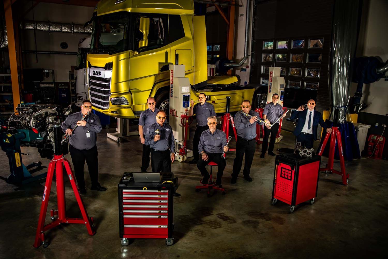 Five musicians standing in a garage with a yellow truck, surrounded by red equipment and stands