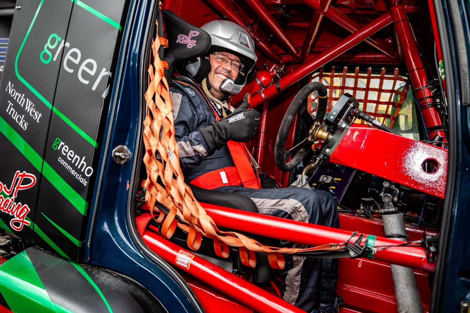 Race car driver seated in a red off-road truck, wearing a helmet and harness inside the roll cage