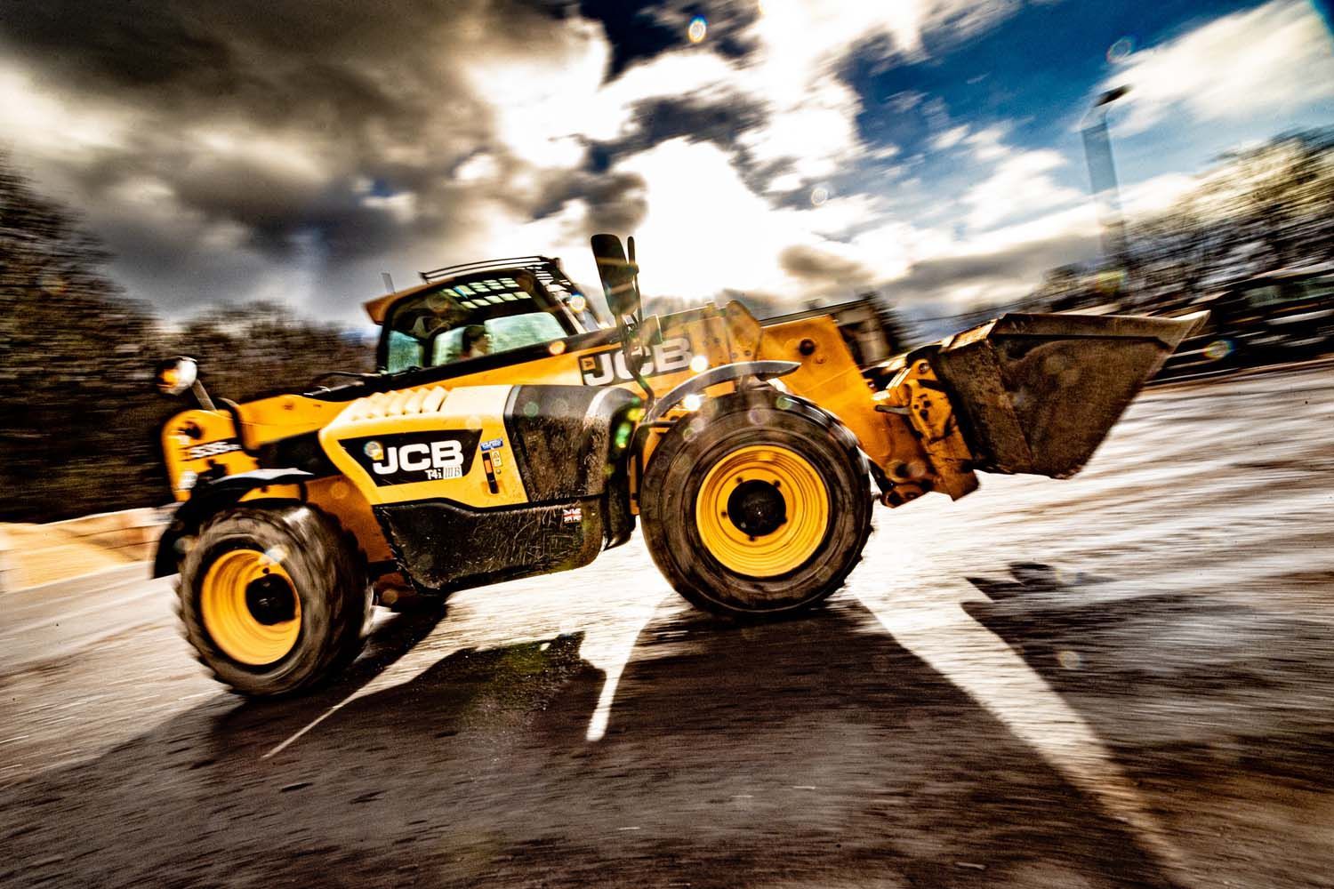 Yellow JCB loader speeding on a dirt road, kicking up dust under a dramatic sky